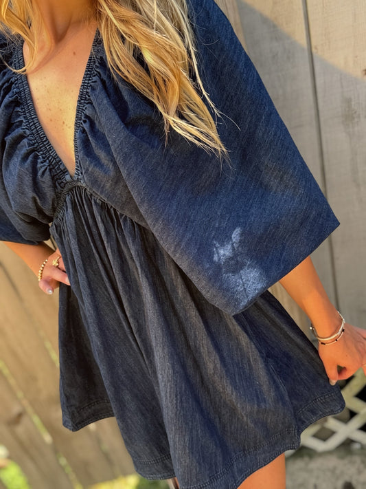 Woman wearing a dark wash denim mini dress with a deep V-neckline, three-quarter puff sleeves and a flared skirt.