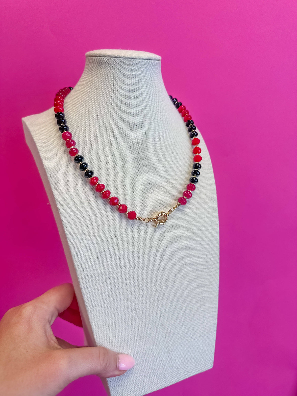 A beaded necklace with alternating red and black beads and a gold clasp, displayed on a beige fabric jewelry bust.