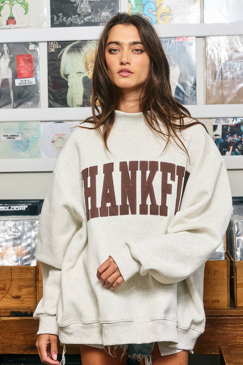 Model wearing a light heathered oversized mock neck sweatshirt with brown THANKFUL appliqué lettering, dropped shoulders, and ribbed trim.