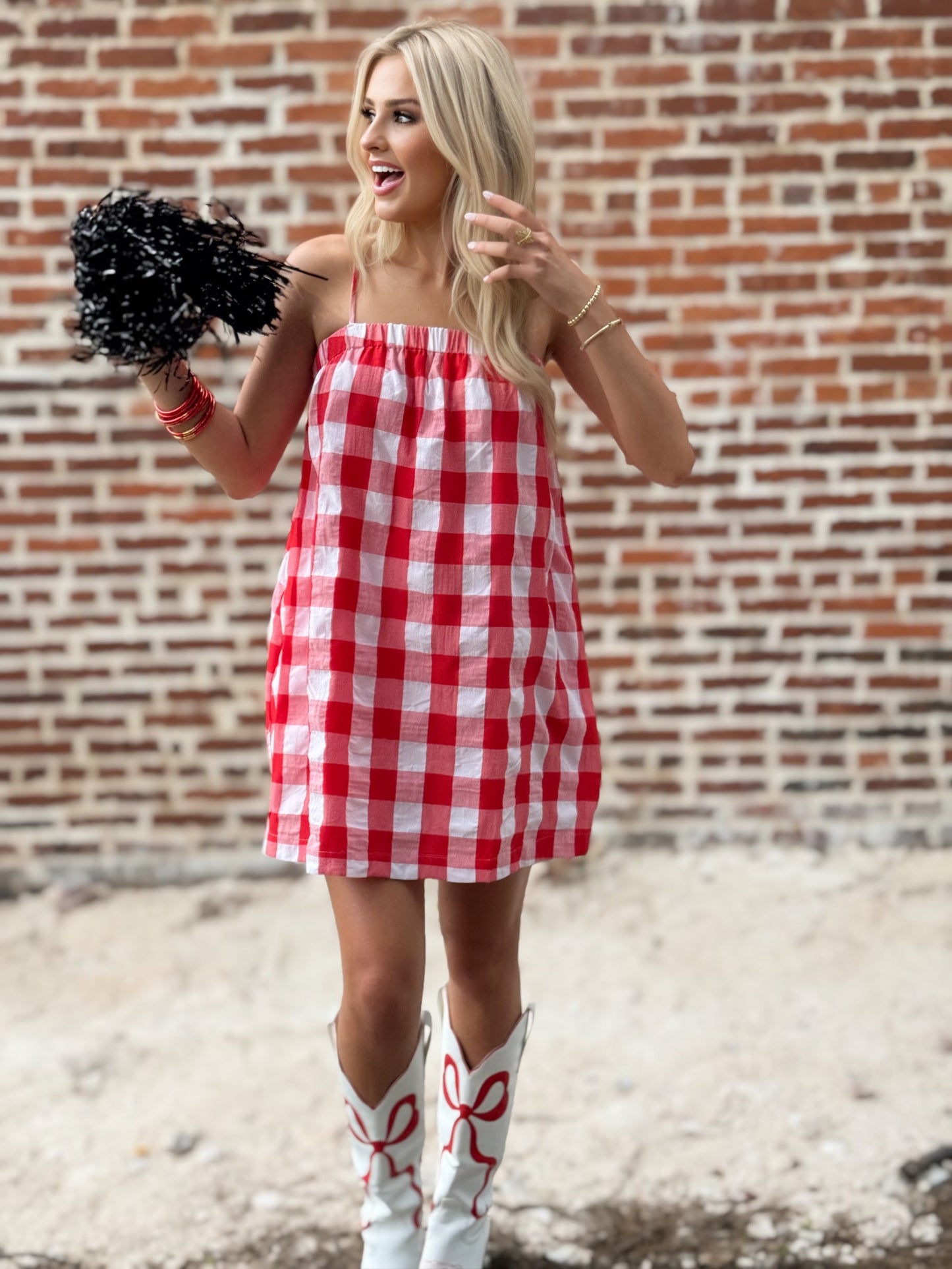 Woman wearing a relaxed red-and-white gingham cami mini dress with adjustable spaghetti straps and hidden side pockets, styled with white cowboy boots.