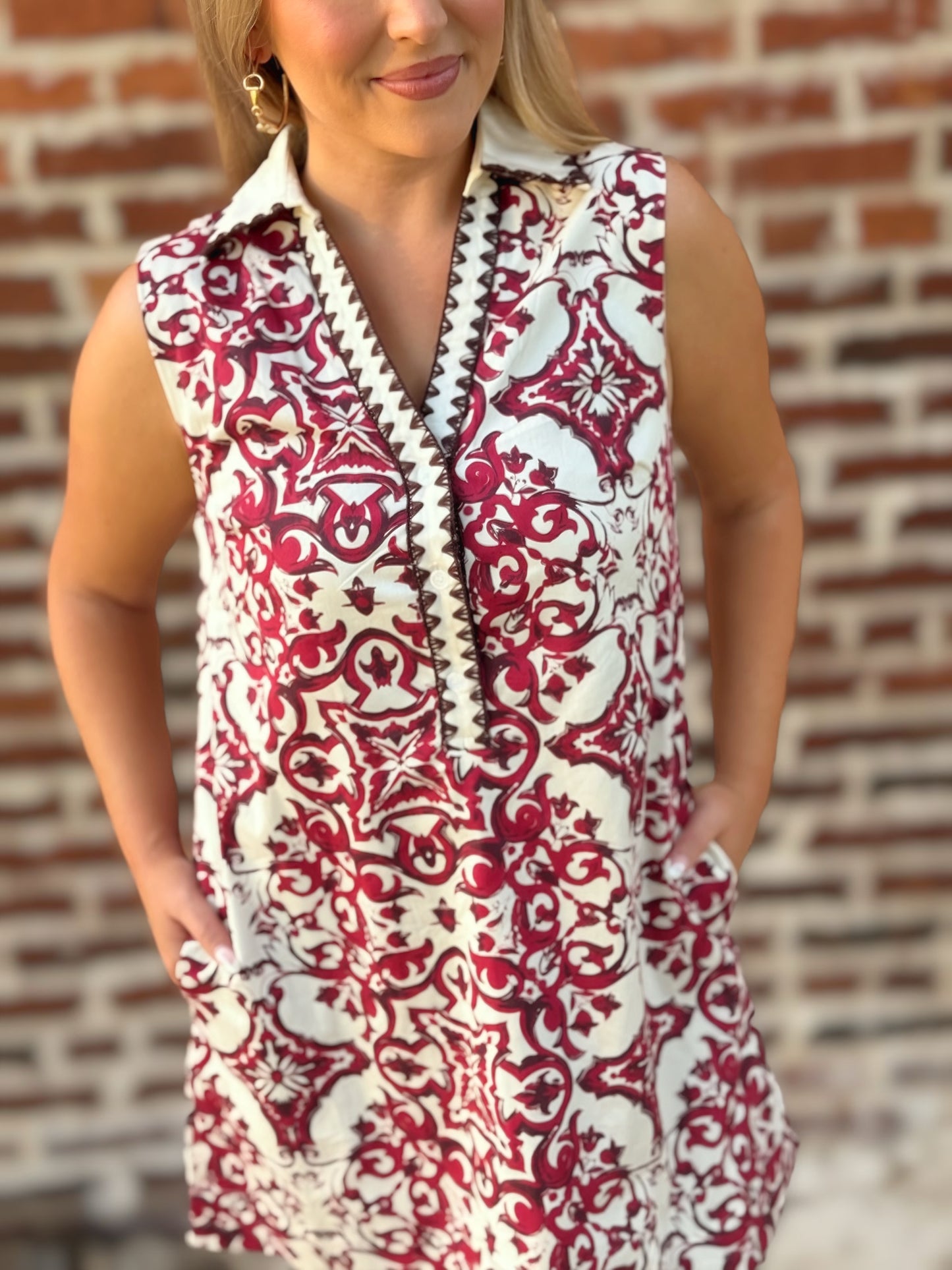 Woman wearing a sleeveless crimson and ivory tile print mini shift dress with a contrast collar, split V-neckline, decorative trim, and hidden pockets.