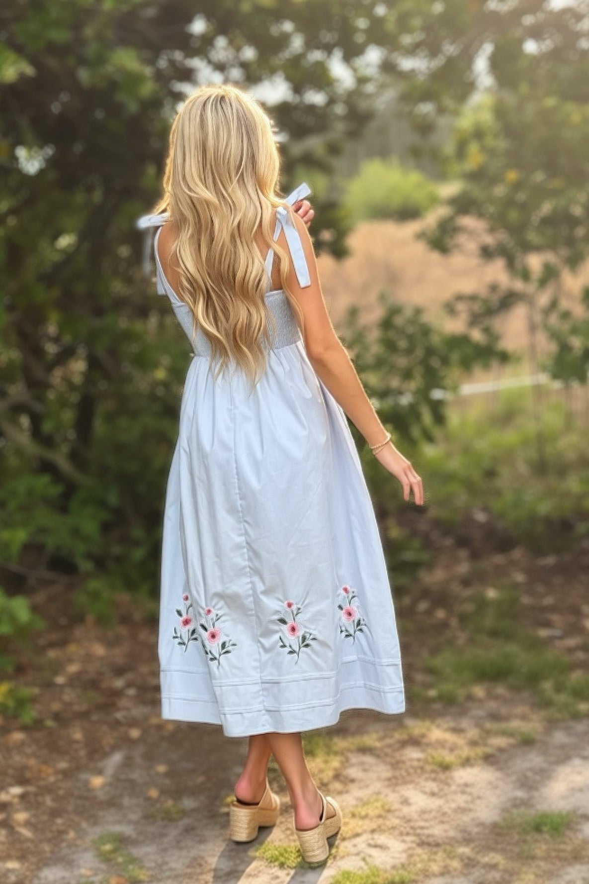 Woman in a light blue dress with floral embroidery walking on a dirt path in a forest.