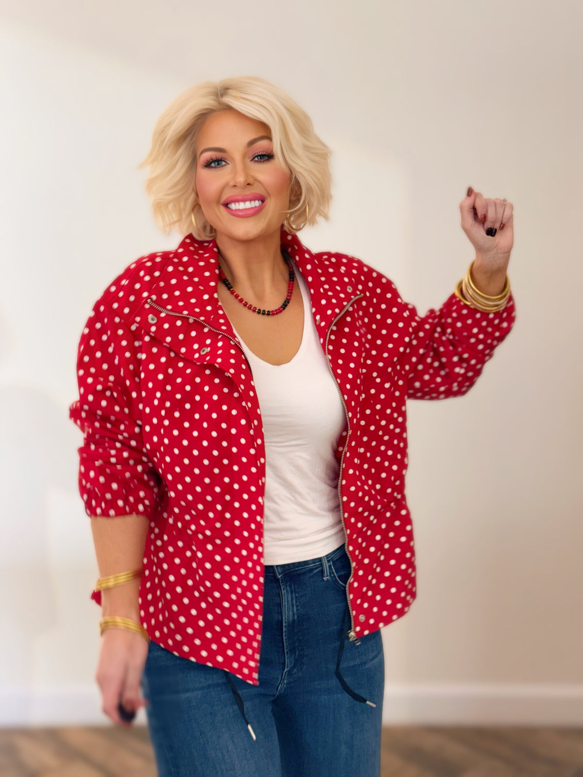 Woman wearing a red and white polka dot jacket with a stand collar, gathered waist, and long sleeves, styled with blue jeans.