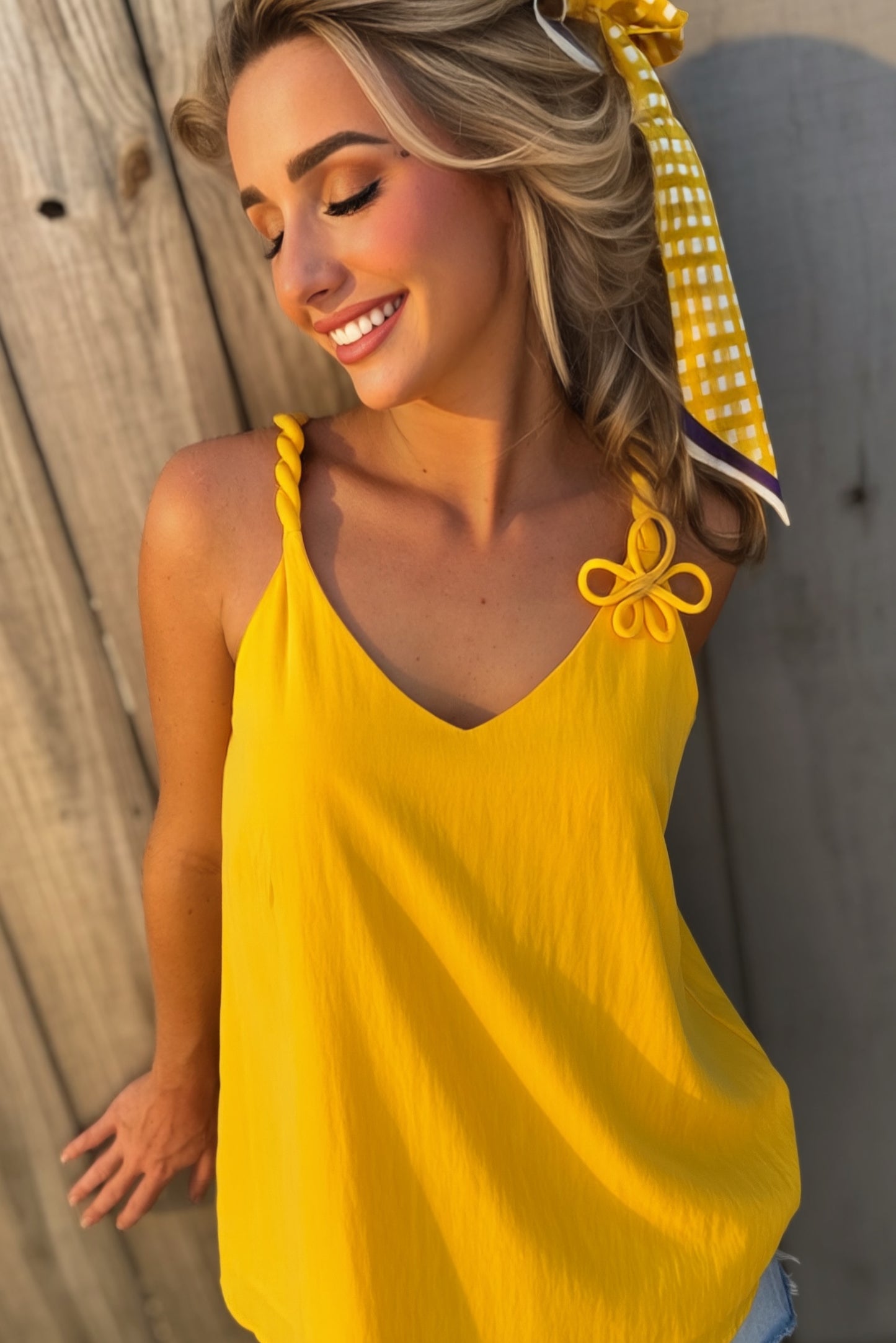 Woman wearing a golden yellow V-neck tank top with twisted cord straps and a sculptural flower detail at the shoulder.