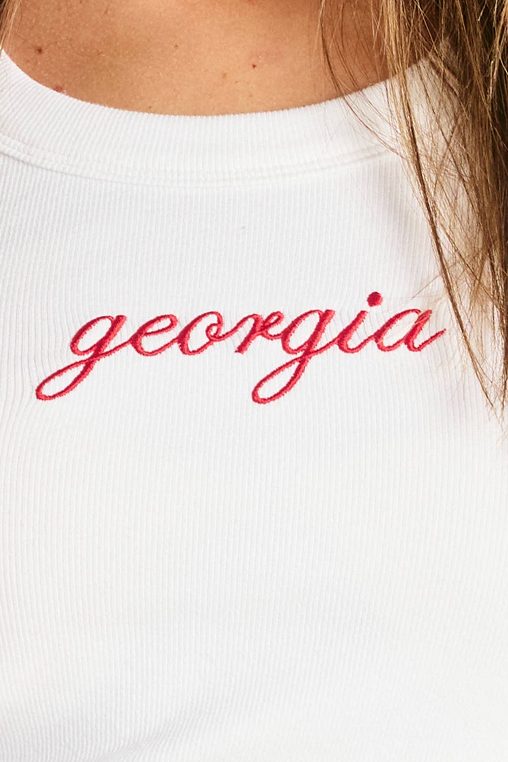 Woman wearing a fitted white ribbed tank top with red cursive “georgia” embroidery.