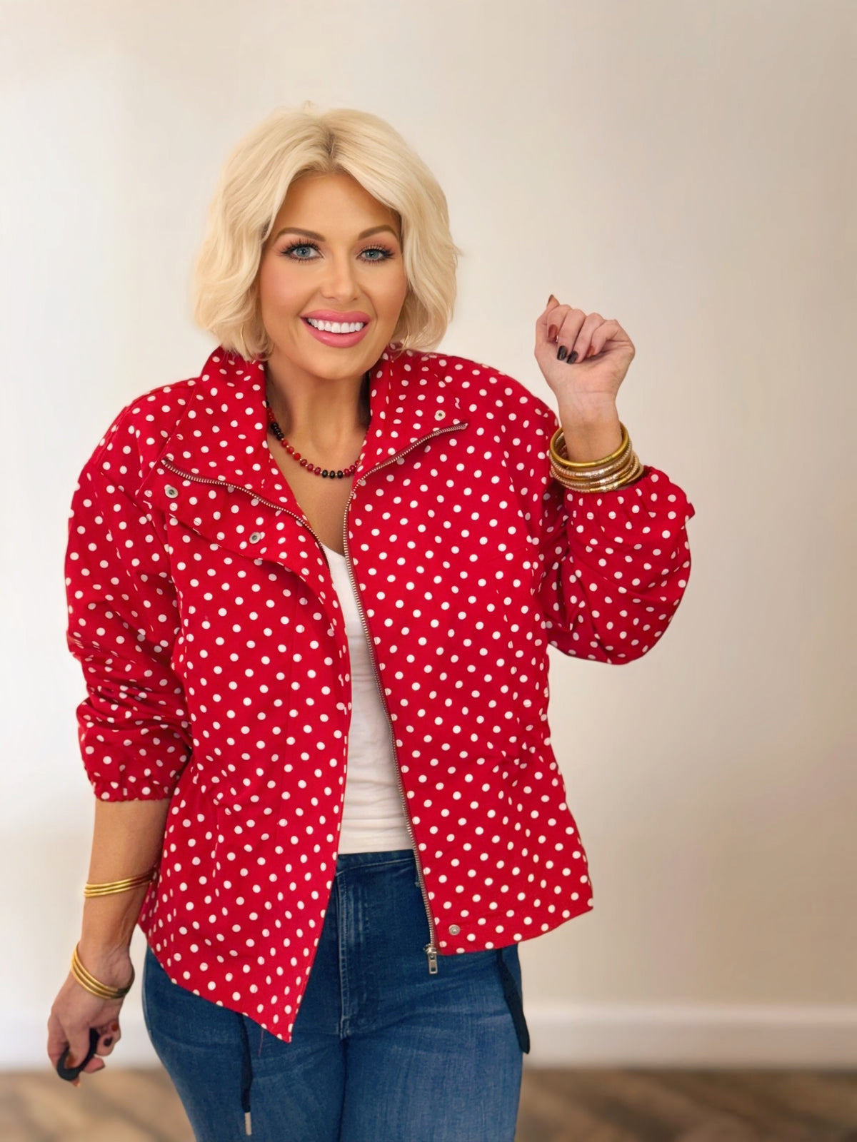 Woman wearing a red and white polka dot jacket with a stand collar, gathered waist, and long sleeves, styled with blue jeans.