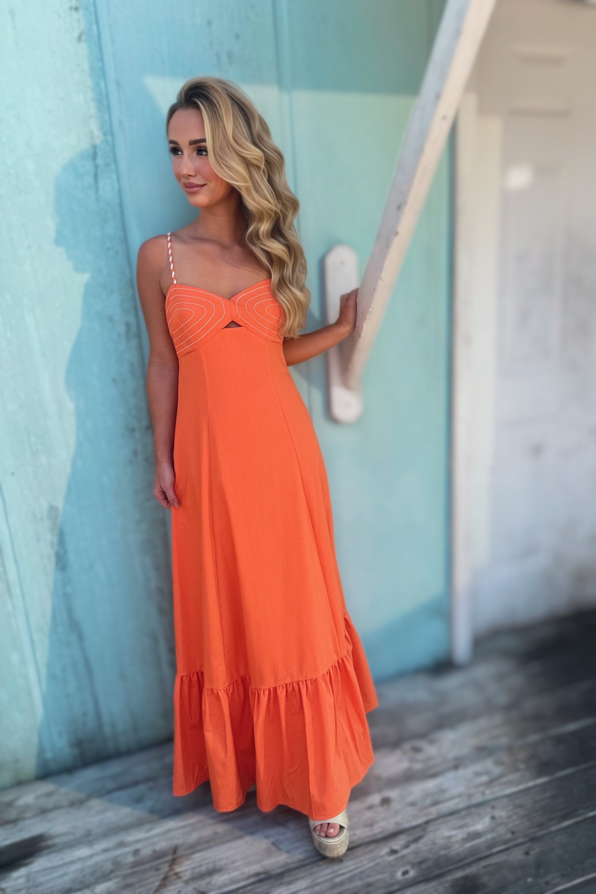 Woman wearing an orange maxi dress with a corset-inspired stitched bodice, braided shoulder straps and a ruffled hem.