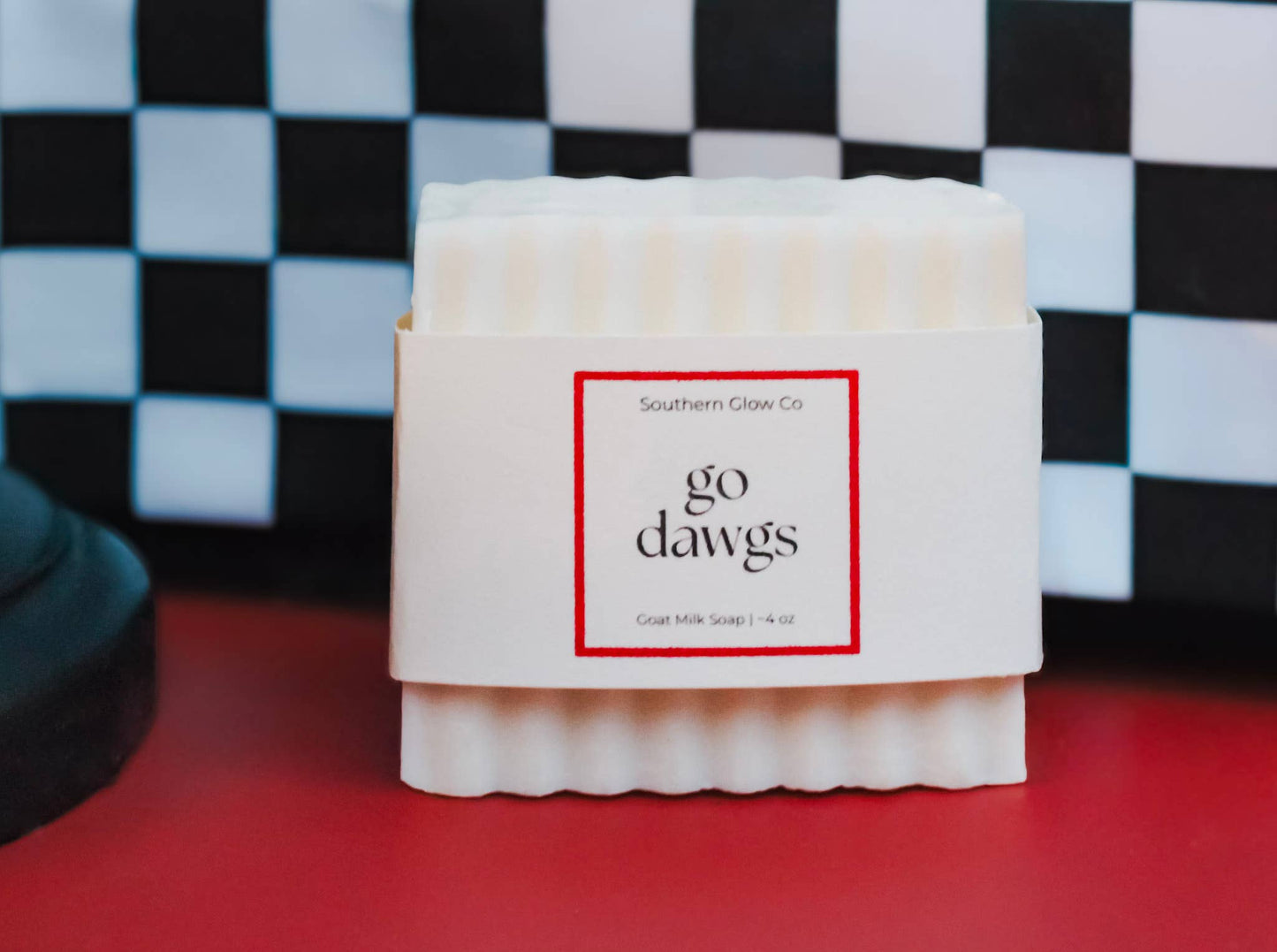 Go Dawgs four-ounce white goat milk soap bar wrapped in red, black and white packaging against a checkered game-day background.