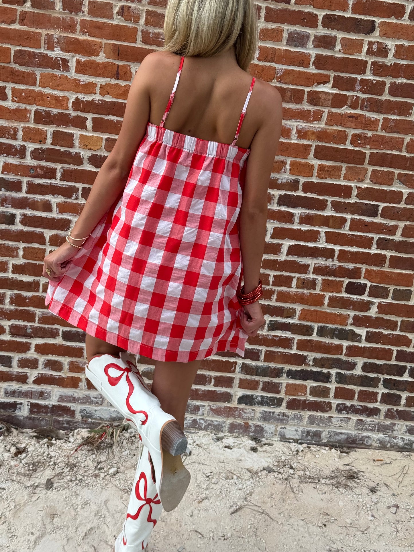 Woman wearing a relaxed red-and-white gingham cami mini dress with adjustable spaghetti straps and hidden side pockets, styled with white cowboy boots.