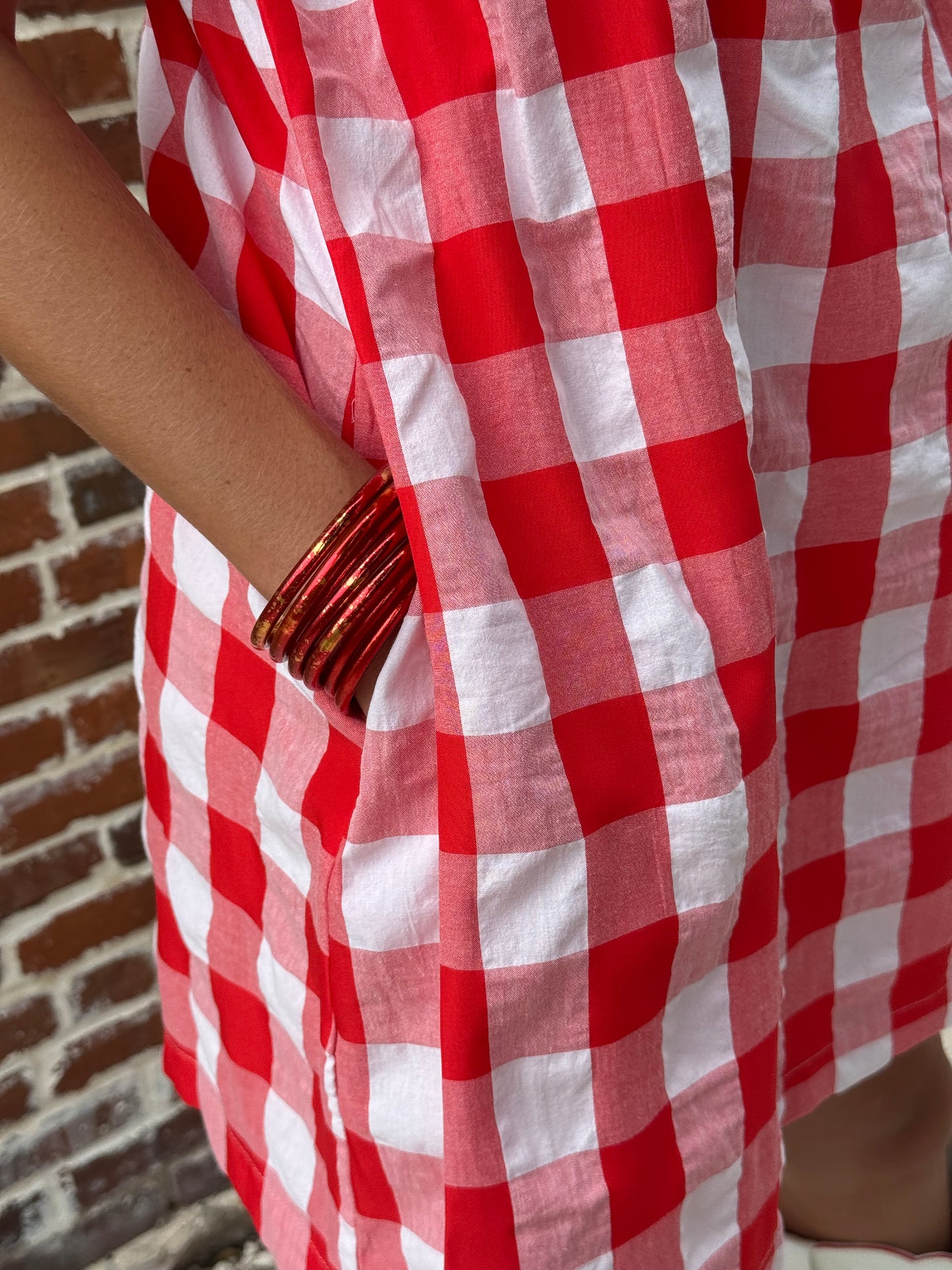 A close-up of a red and white checkered fabric, likely a skirt or drape, being held by a person wearing gold bangles. A brick floor is visible in the background.