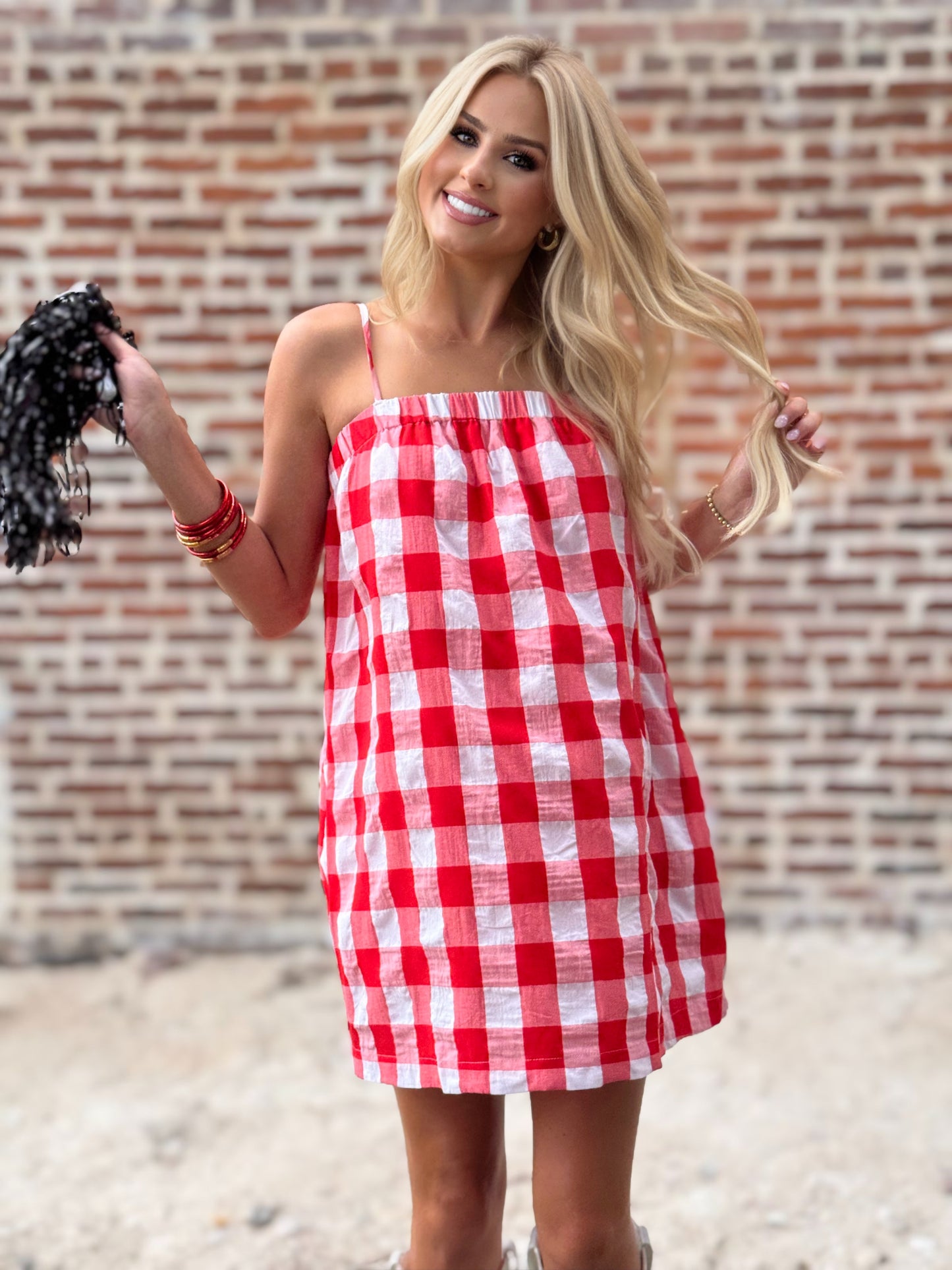 Woman wearing a relaxed red-and-white gingham cami mini dress with adjustable spaghetti straps and hidden side pockets, styled with white cowboy boots.
