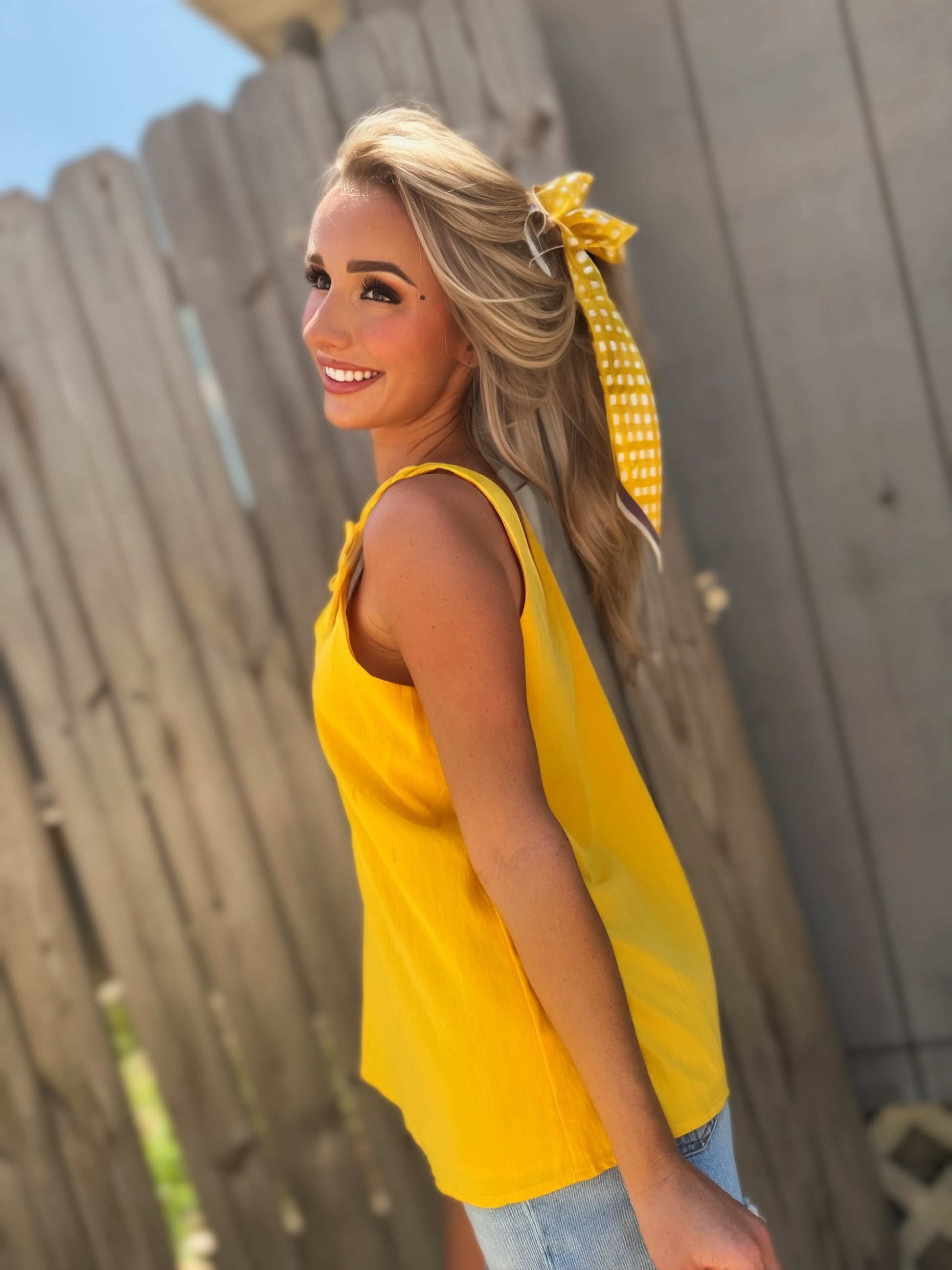 Woman wearing a golden yellow V-neck tank top with twisted cord straps and a sculptural flower detail at the shoulder.