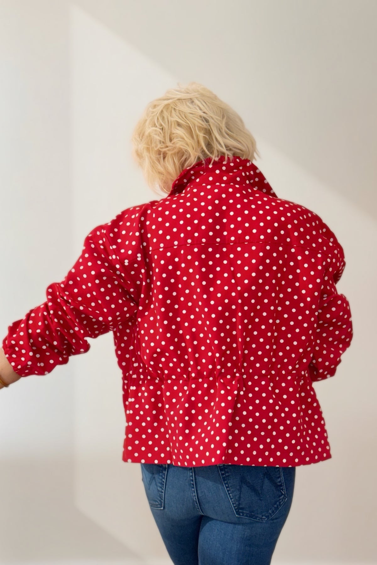 Woman wearing a red and white polka dot jacket with a stand collar, gathered waist, and long sleeves, styled with blue jeans.
