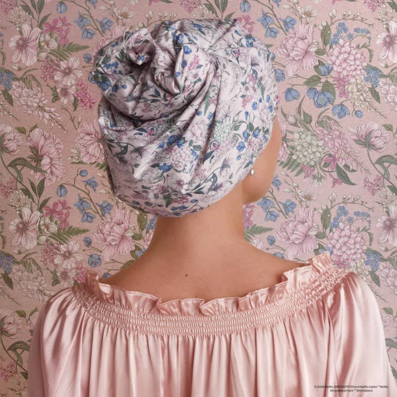 Person wearing a floral headscarf and pink satin nightgown against a floral wallpaper background