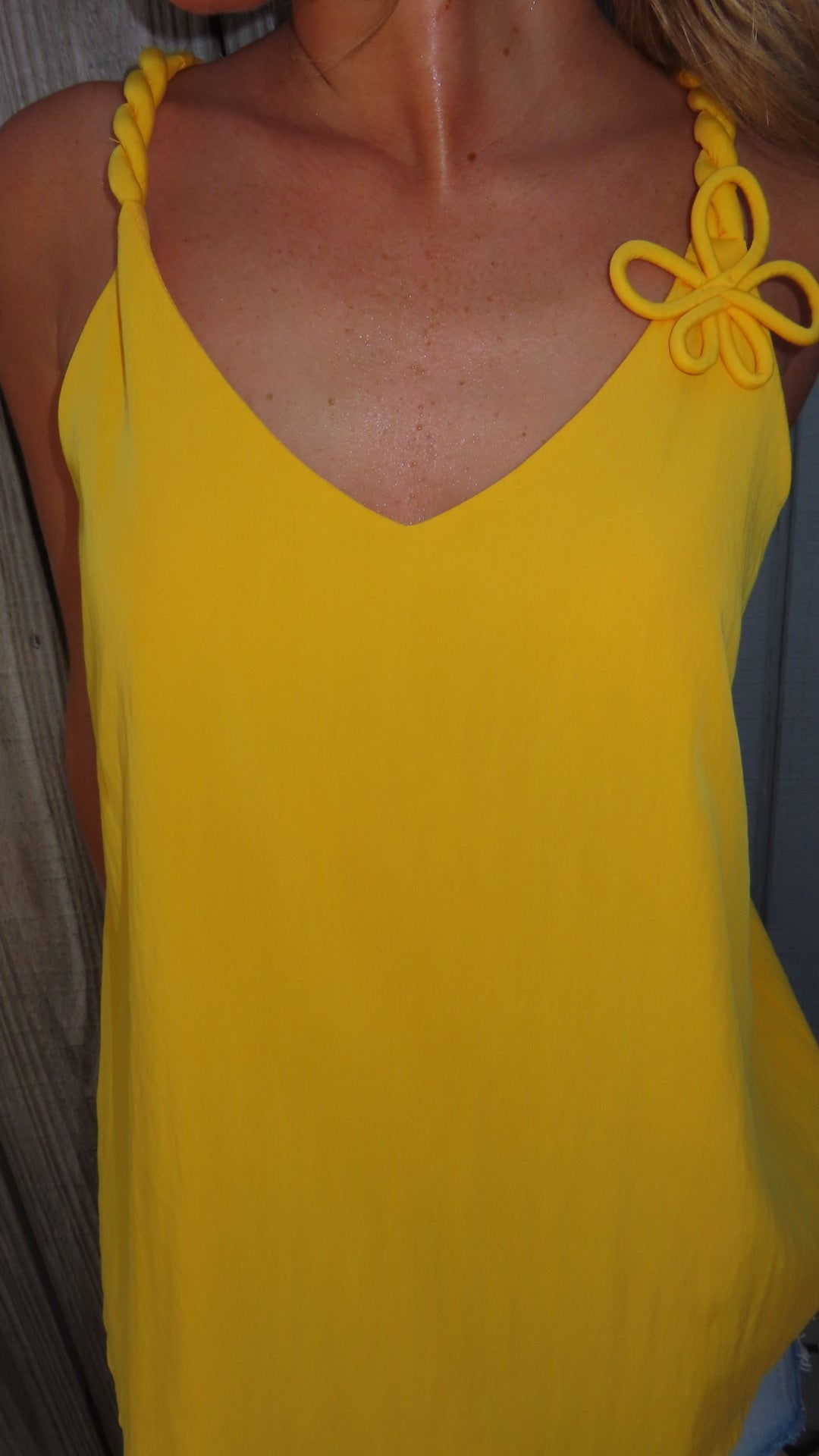 Woman wearing a golden yellow V-neck tank top with twisted cord straps and a sculptural flower detail at the shoulder.