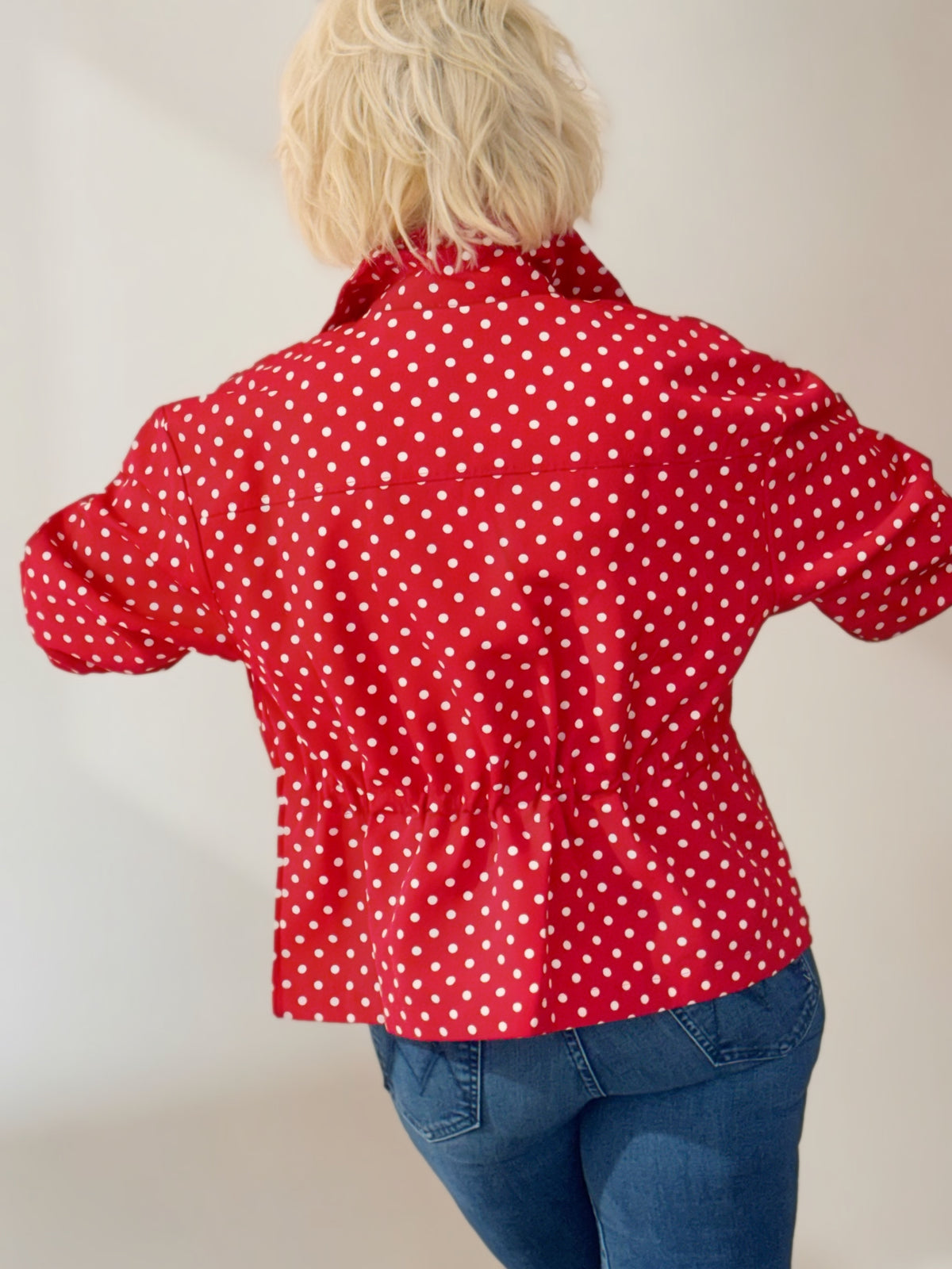 Woman wearing a red and white polka dot jacket with a stand collar, gathered waist, and long sleeves, styled with blue jeans.