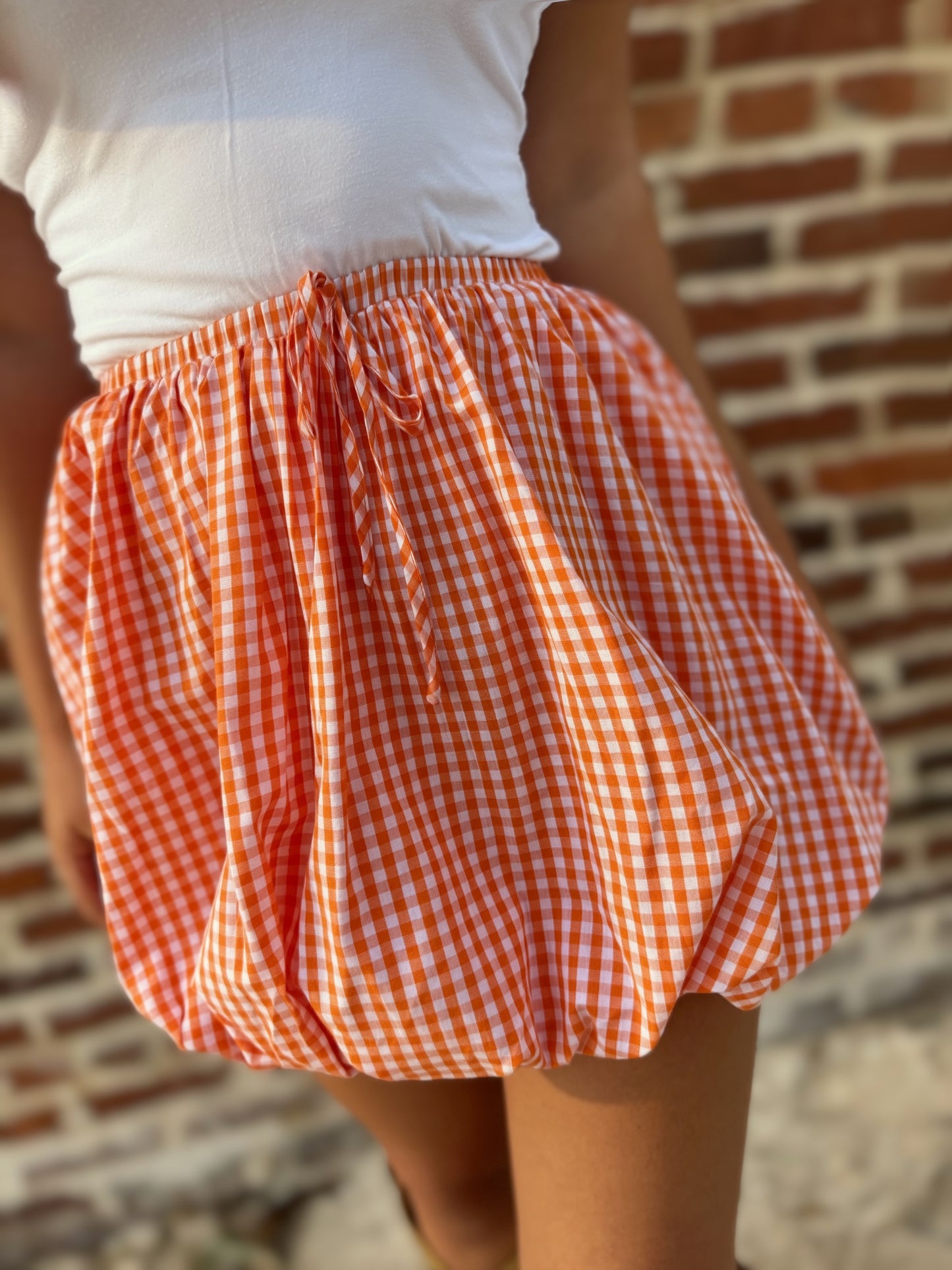 A close-up of a person wearing a high-waisted, voluminous orange and white gingham skirt with a drawstring waist, paired with a white top, set against a blurred brick wall background.