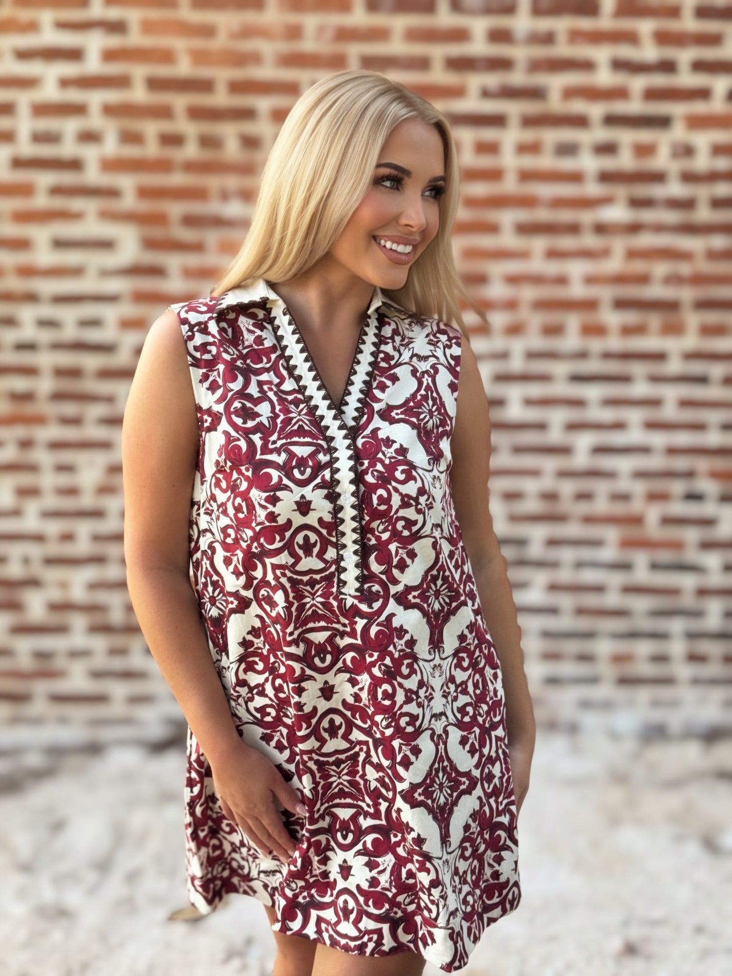 Woman wearing a sleeveless crimson and ivory tile print mini shift dress with a contrast collar, split V-neckline, decorative trim, and hidden pockets.