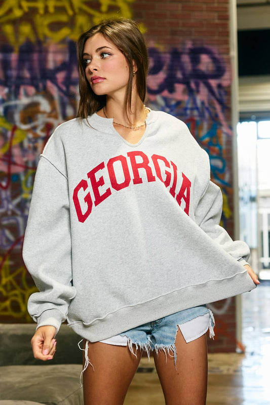 Woman wearing an oversized heather grey split-neck sweatshirt with large red Georgia lettering, drop shoulders and a front pocket.