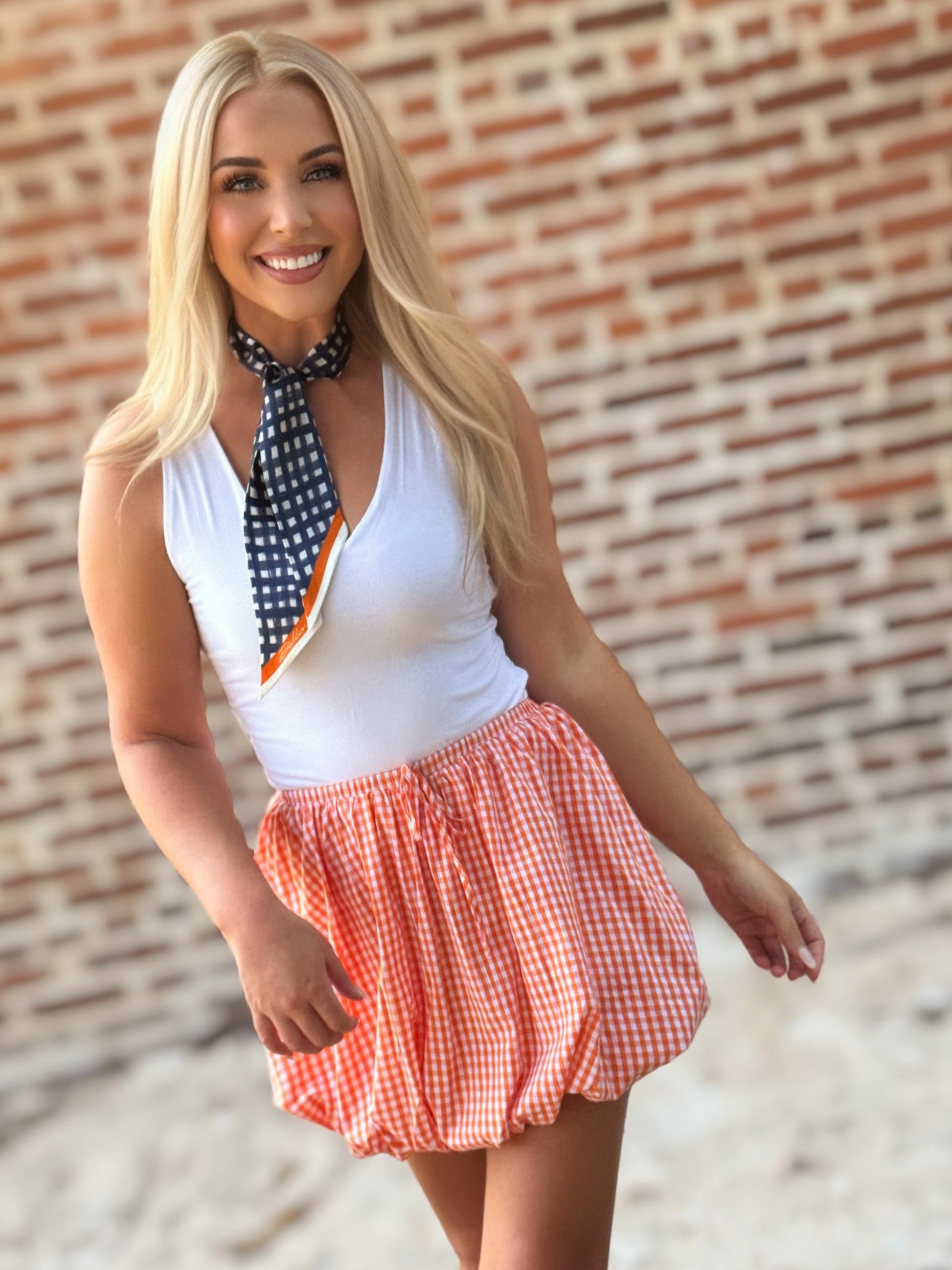 Woman wearing an orange-and-white gingham bubble mini skirt styled with a white sleeveless top, scarf and cowboy boots.