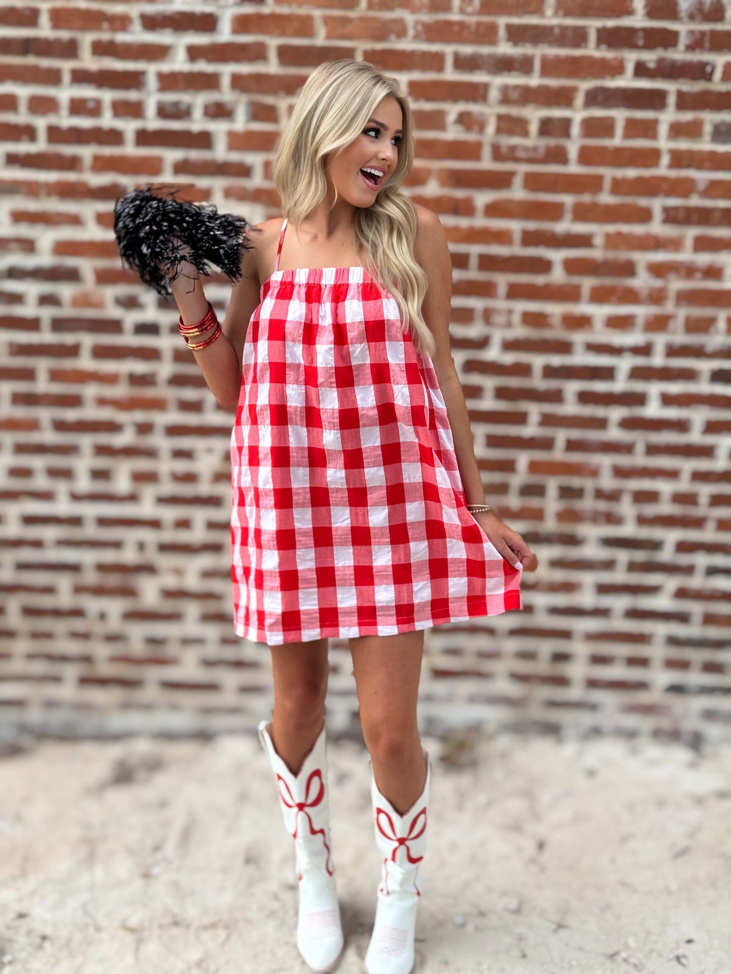 Woman wearing a relaxed red-and-white gingham cami mini dress with adjustable spaghetti straps and hidden side pockets, styled with white cowboy boots.