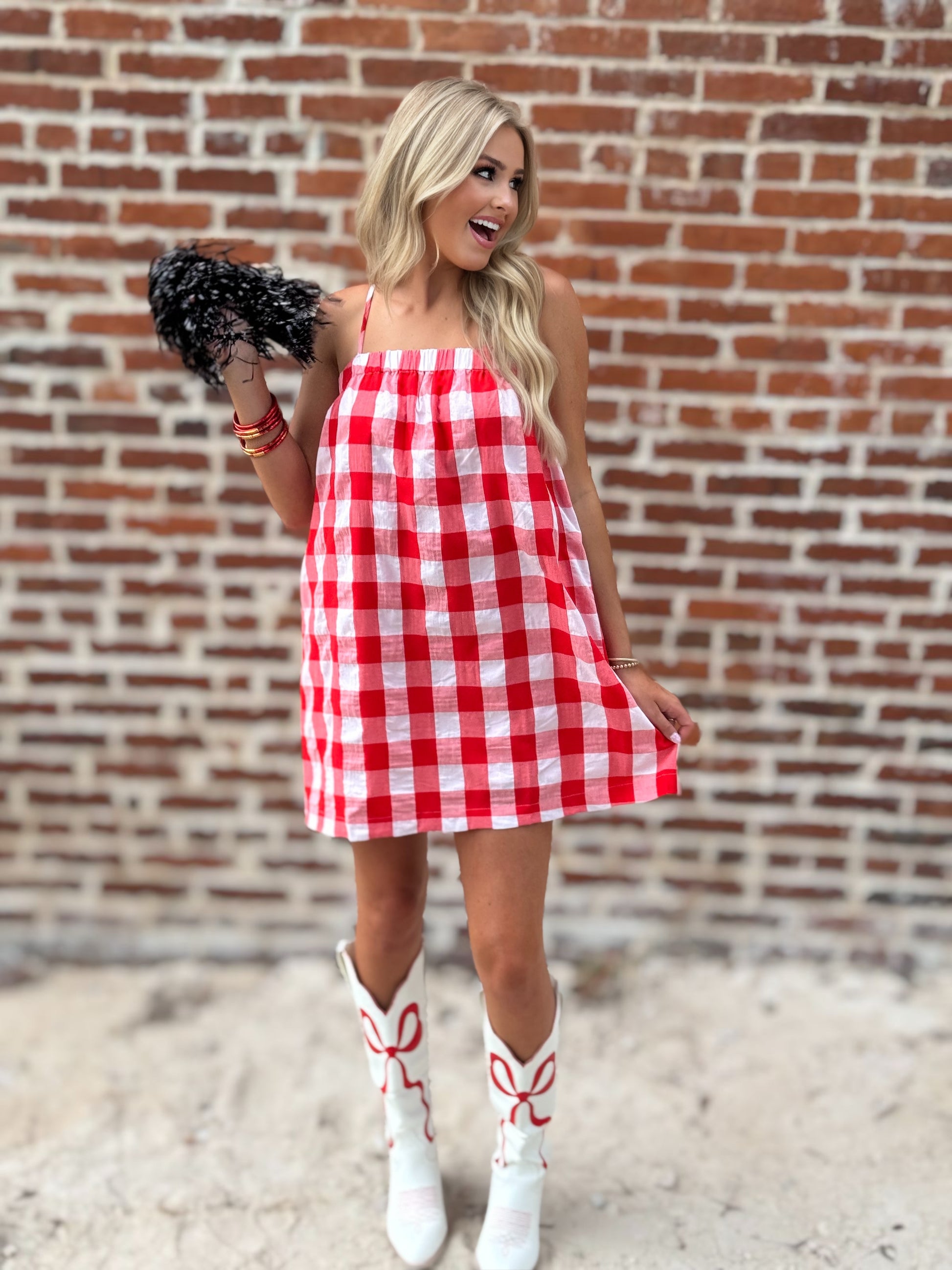 Woman wearing a relaxed red-and-white gingham cami mini dress with adjustable spaghetti straps and hidden side pockets, styled with white cowboy boots.