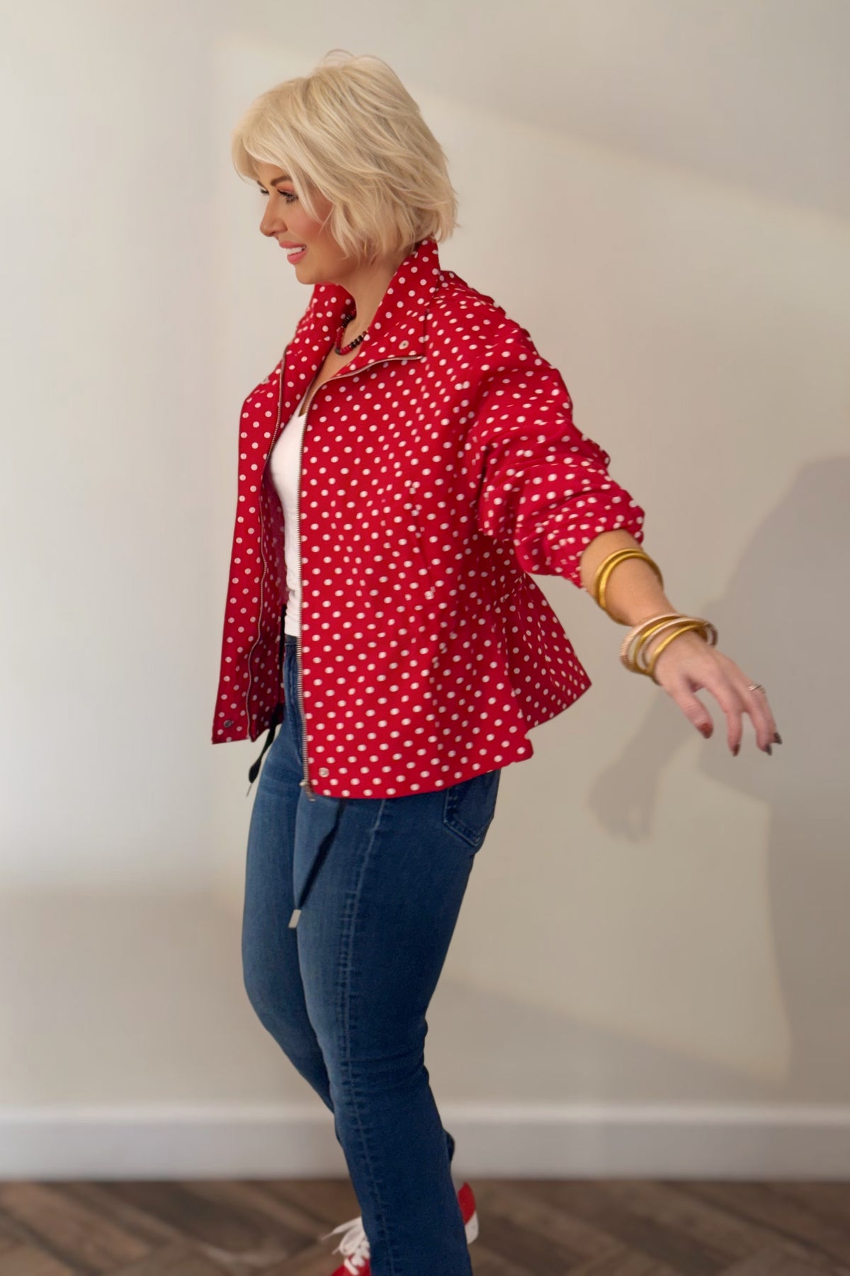 Woman wearing a red and white polka dot jacket with a stand collar, gathered waist, and long sleeves, styled with blue jeans.