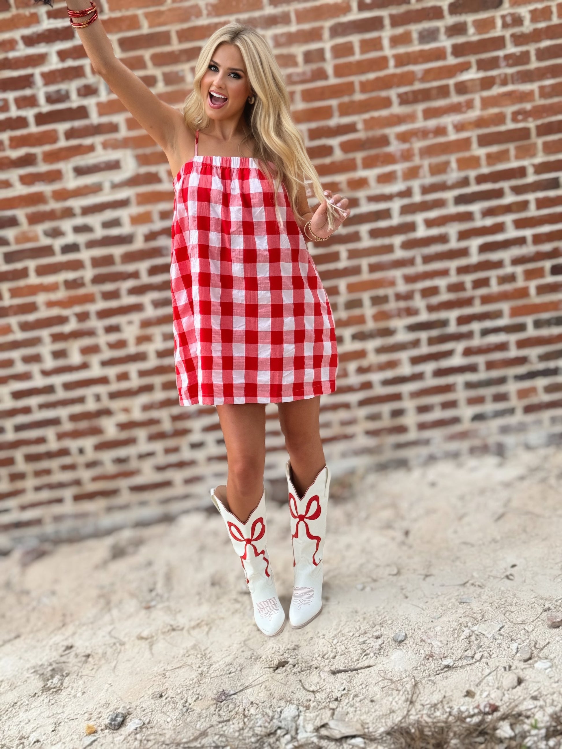 Woman wearing a relaxed red-and-white gingham cami mini dress with adjustable spaghetti straps and hidden side pockets, styled with white cowboy boots.