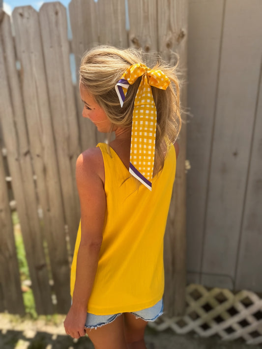 Woman wearing a golden yellow V-neck tank top with twisted cord straps and a sculptural flower detail at the shoulder.