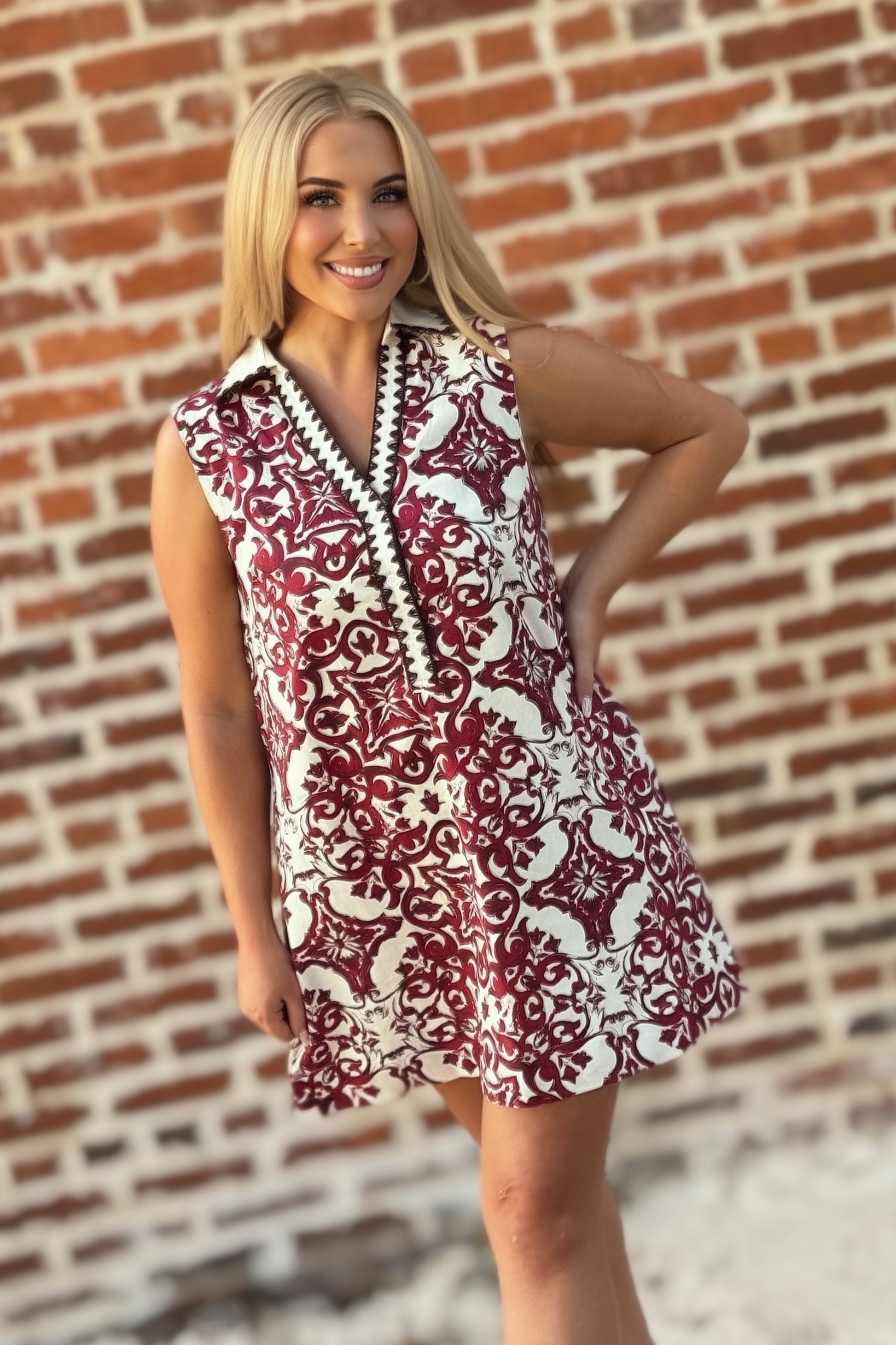 Woman wearing a sleeveless crimson and ivory tile print mini shift dress with a contrast collar, split V-neckline, decorative trim, and hidden pockets.
