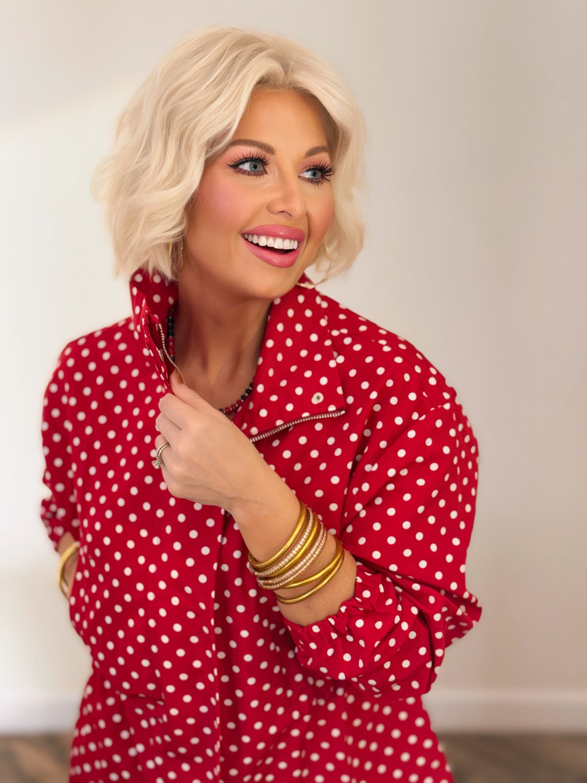 Woman wearing a red and white polka dot jacket with a stand collar, gathered waist, and long sleeves, styled with blue jeans.