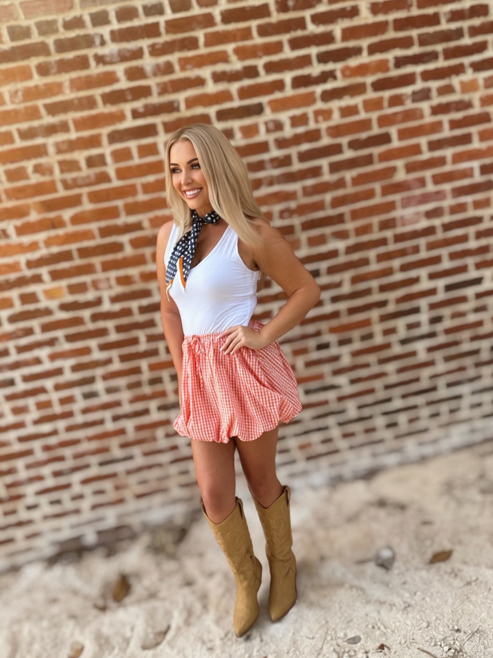 Woman wearing an orange-and-white gingham bubble mini skirt styled with a white sleeveless top, scarf and cowboy boots.
