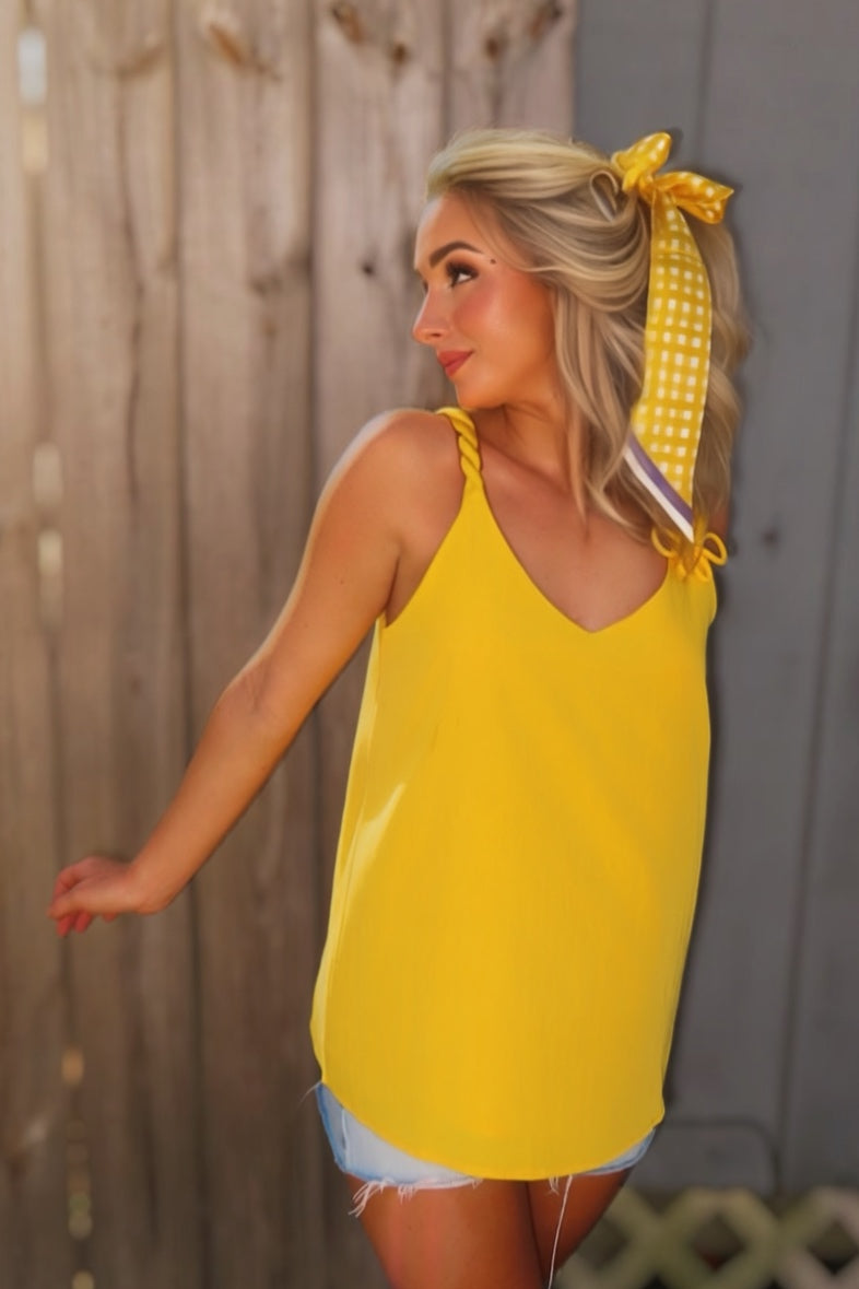 Woman wearing a golden yellow V-neck tank top with twisted cord straps and a sculptural flower detail at the shoulder.