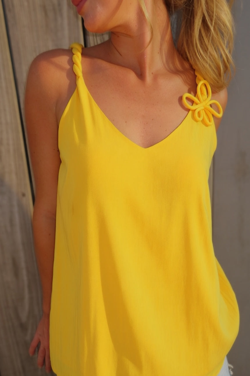 Woman wearing a golden yellow V-neck tank top with twisted cord straps and a sculptural flower detail at the shoulder.