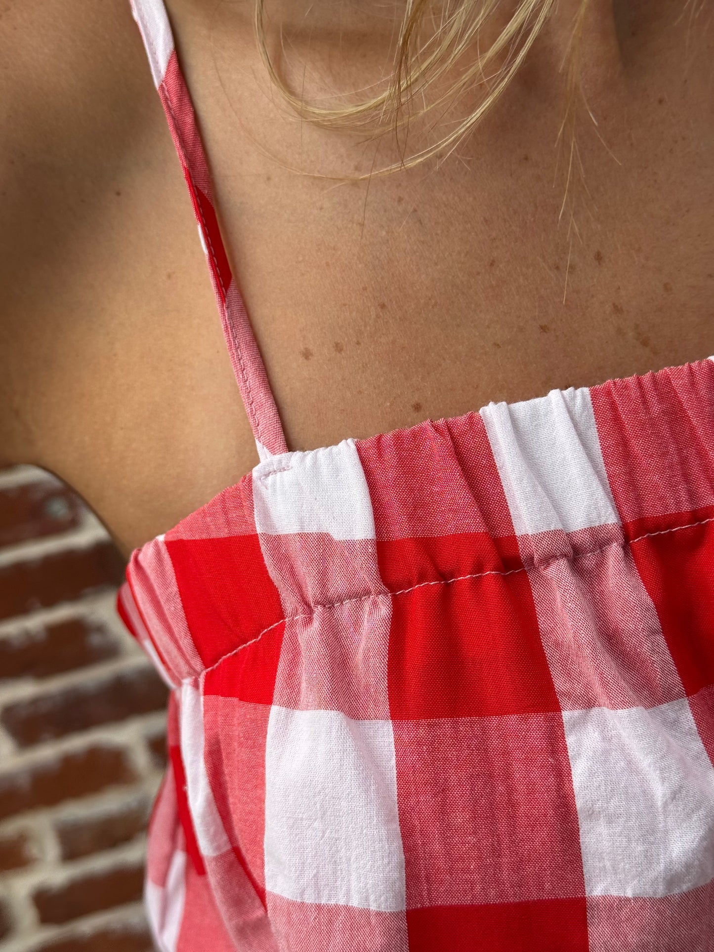 Close-up of a red and white gingham checkered garment with a ruffled edge, worn by a person whose bare shoulder and hair are visible.
