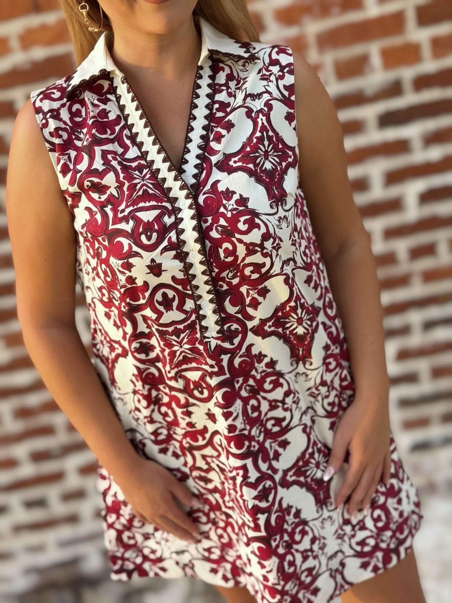 Woman wearing a sleeveless crimson and ivory tile print mini shift dress with a contrast collar, split V-neckline, decorative trim, and hidden pockets.