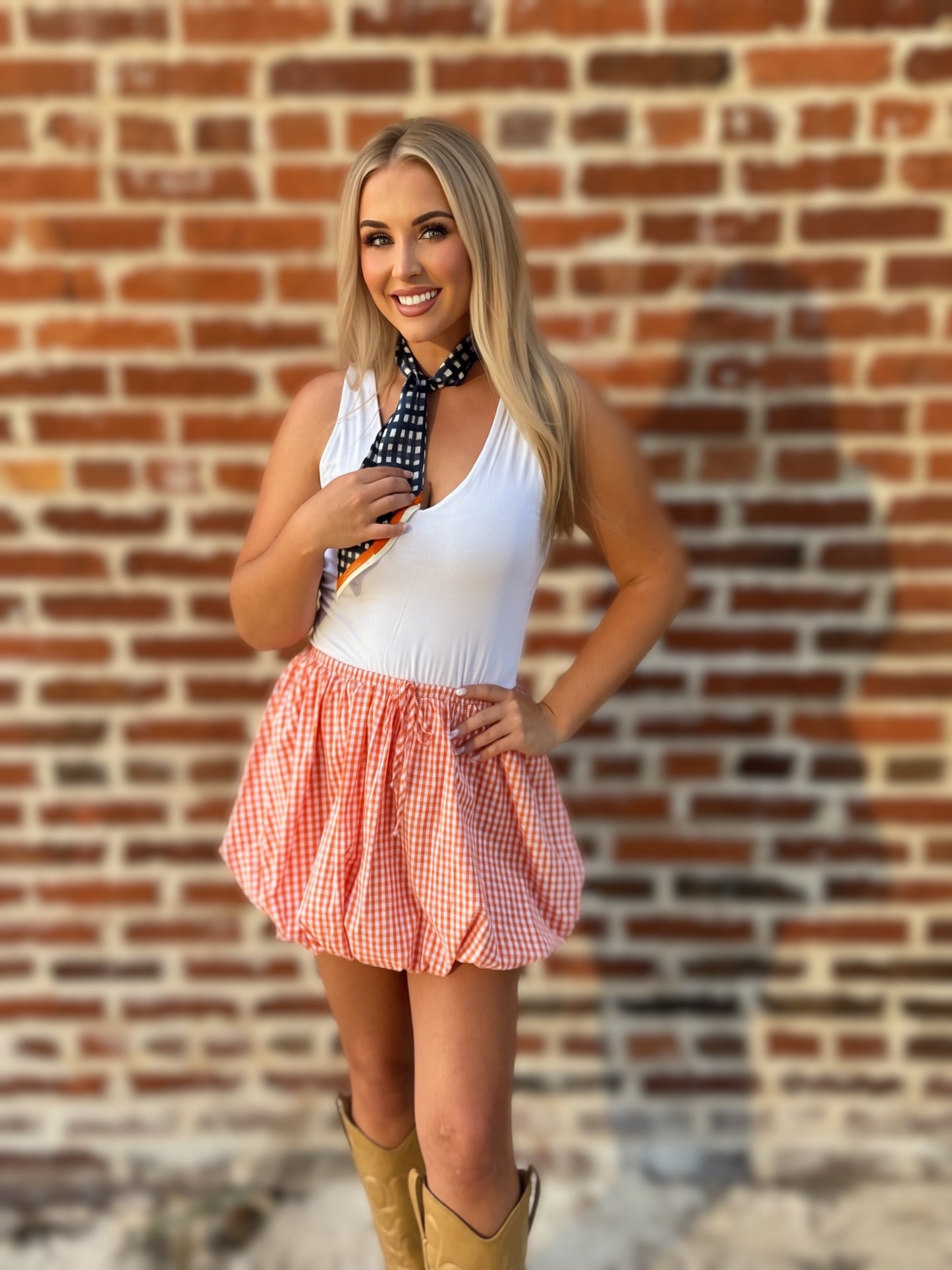 Woman wearing an orange-and-white gingham bubble mini skirt styled with a white sleeveless top, scarf and cowboy boots.