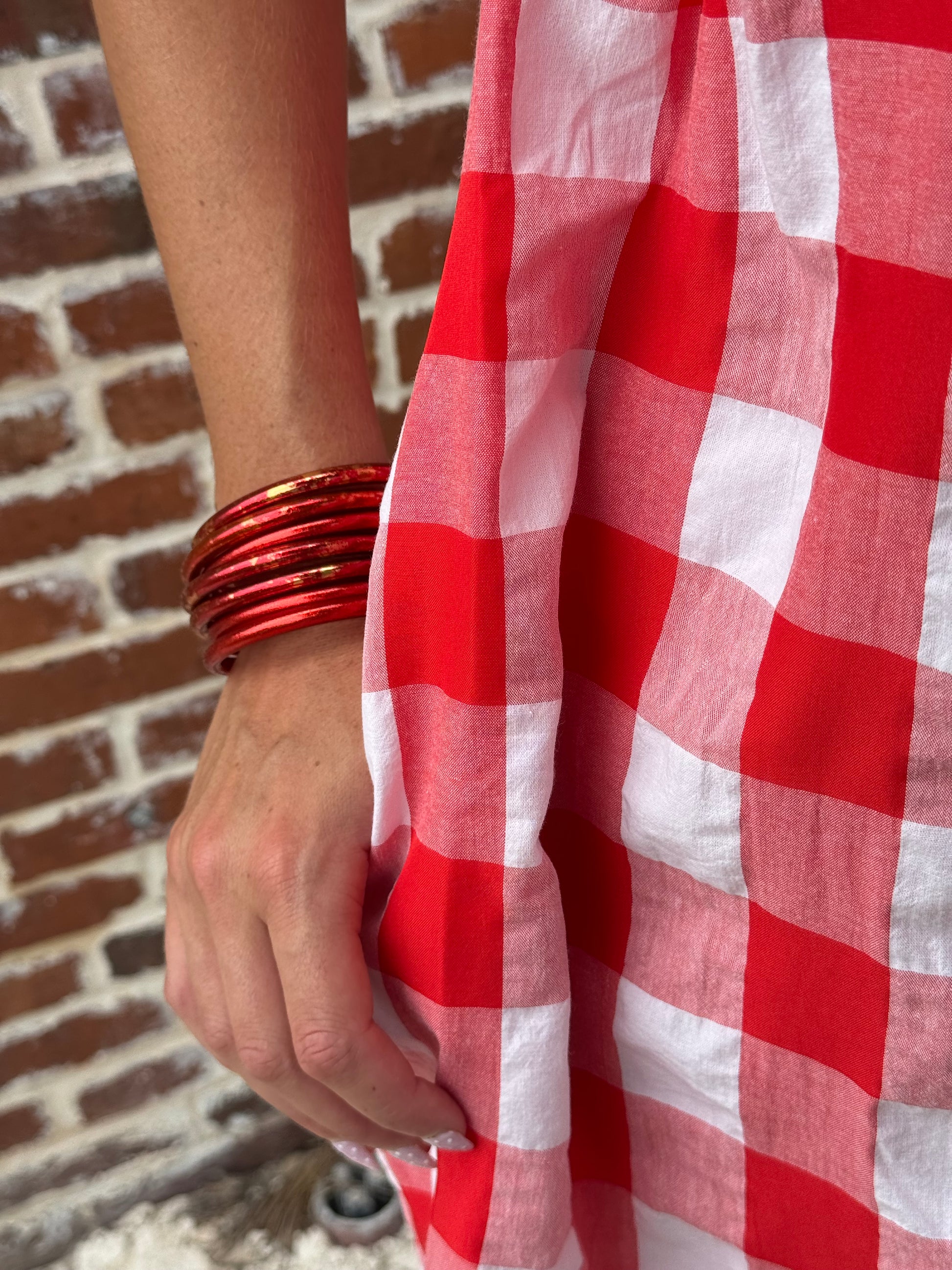 A close-up of a person holding a red and white checkered fabric, wearing red bangles, with a brick wall in the background.