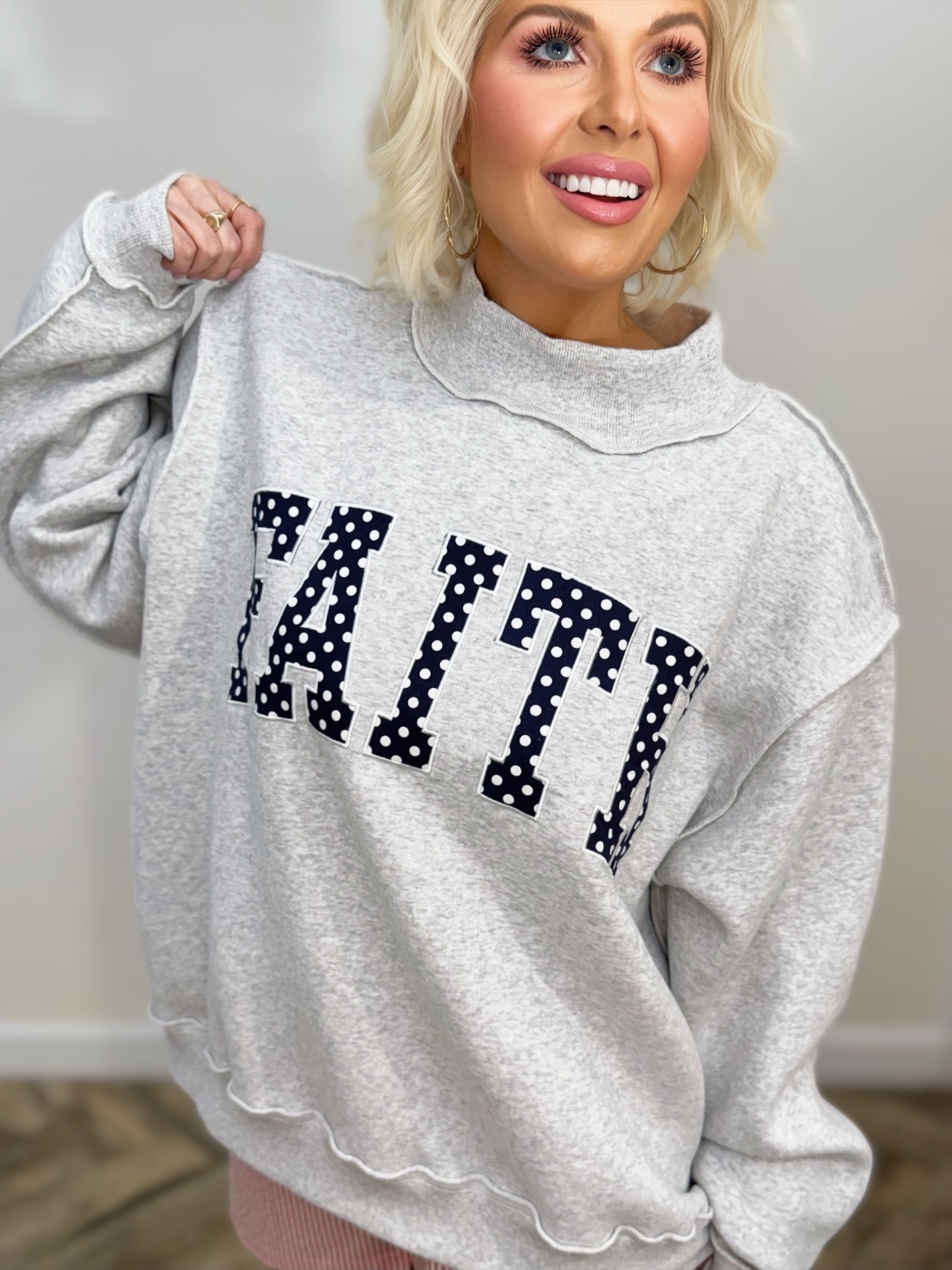 Heather gray oversized mock-neck sweatshirt featuring bold navy polka-dot "FAITH" embroidered lettering on the front