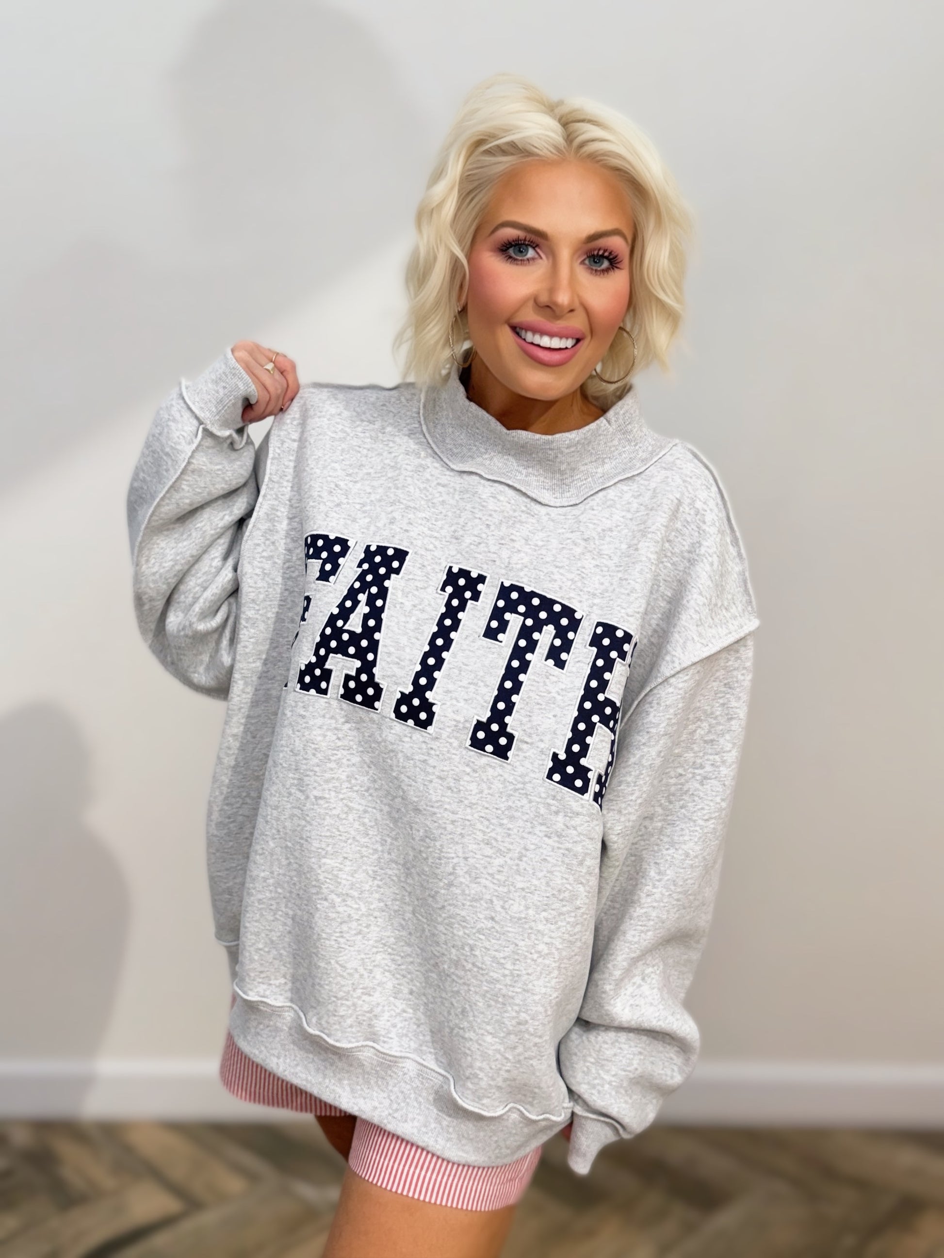 Heather gray oversized mock-neck sweatshirt featuring bold navy polka-dot "FAITH" embroidered lettering on the front