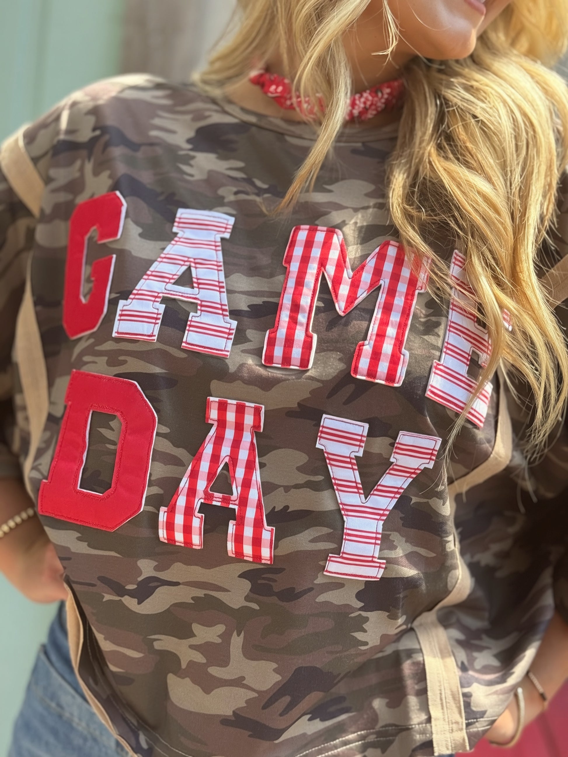 Woman wearing an oversized washed camouflage game day top with red, white and striped patchwork lettering.