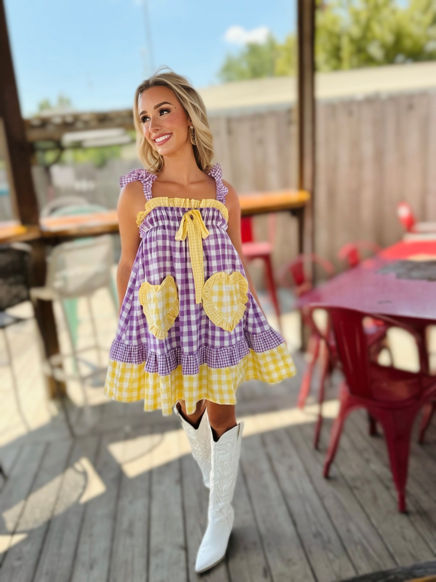 Woman wearing a purple and gold gingham babydoll mini dress with ruffle trim, a front tie and oversized heart-shaped pockets.