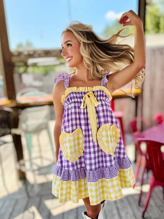 Woman wearing a purple and gold gingham babydoll mini dress with ruffle trim, a front tie and oversized heart-shaped pockets.