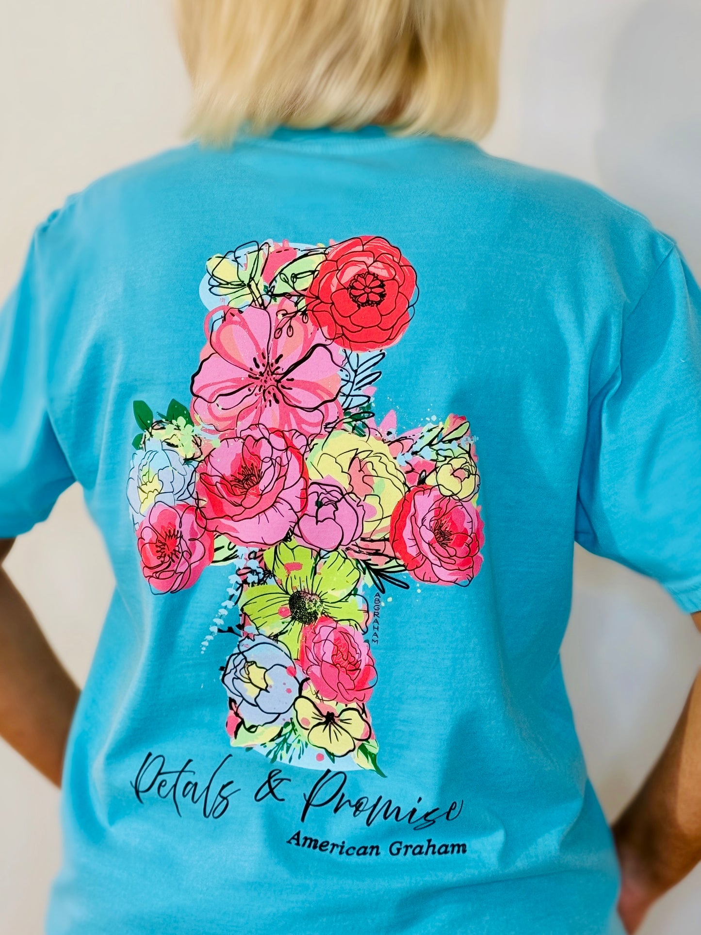 Person wearing a blue t-shirt with colorful floral design and 'Petals & Promise' text.