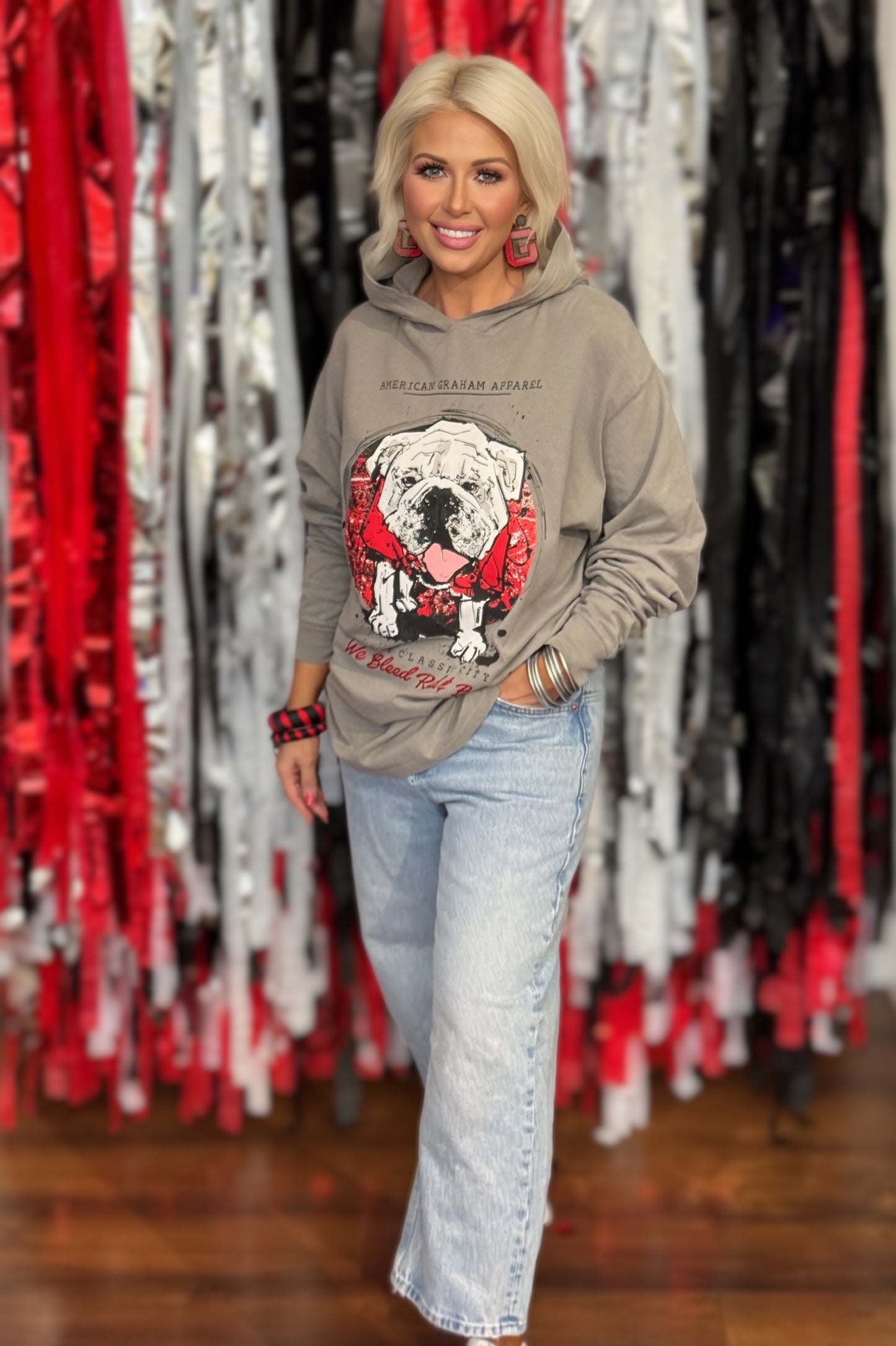 A person wearing a gray hooded long sleeve t-shirt with a large graphic print of a bulldog wearing a red bandana, with the text 'AMERICAN GRAHAM APPAREL' and 'The Classic City We Blood Red & Black' on the front.