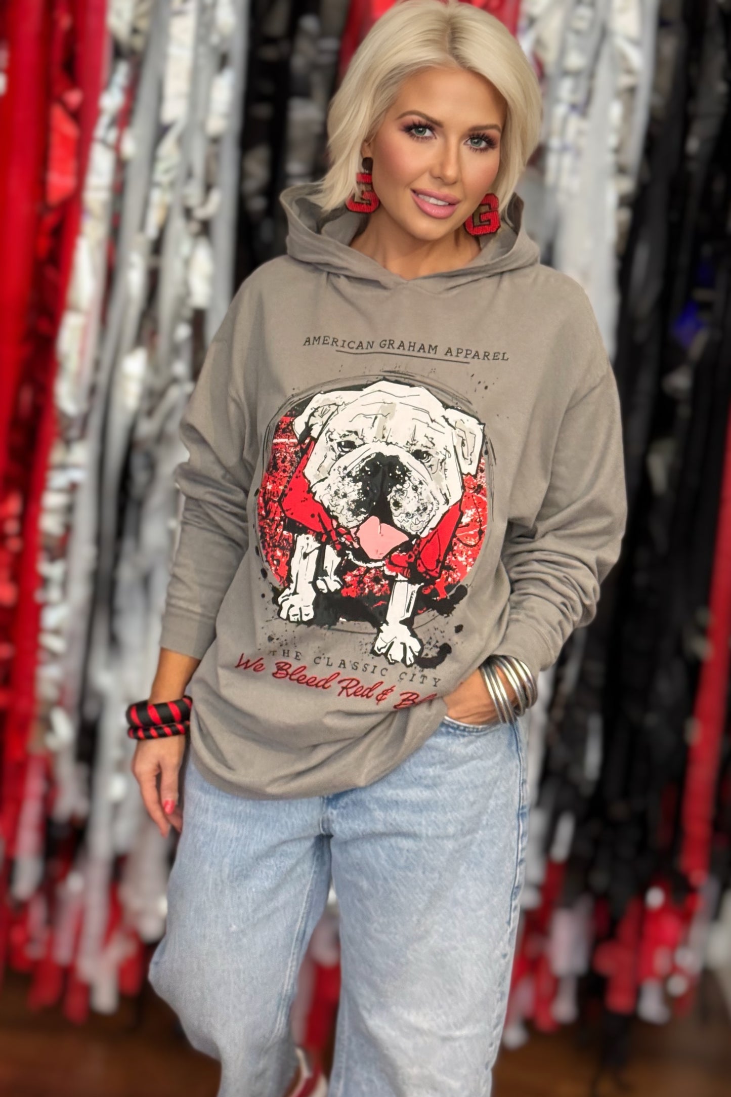 A person wearing a gray hooded long sleeve t-shirt with a large graphic print of a bulldog wearing a red bandana, with the text 'AMERICAN GRAHAM APPAREL' and 'The Classic City We Blood Red & Black' on the front.