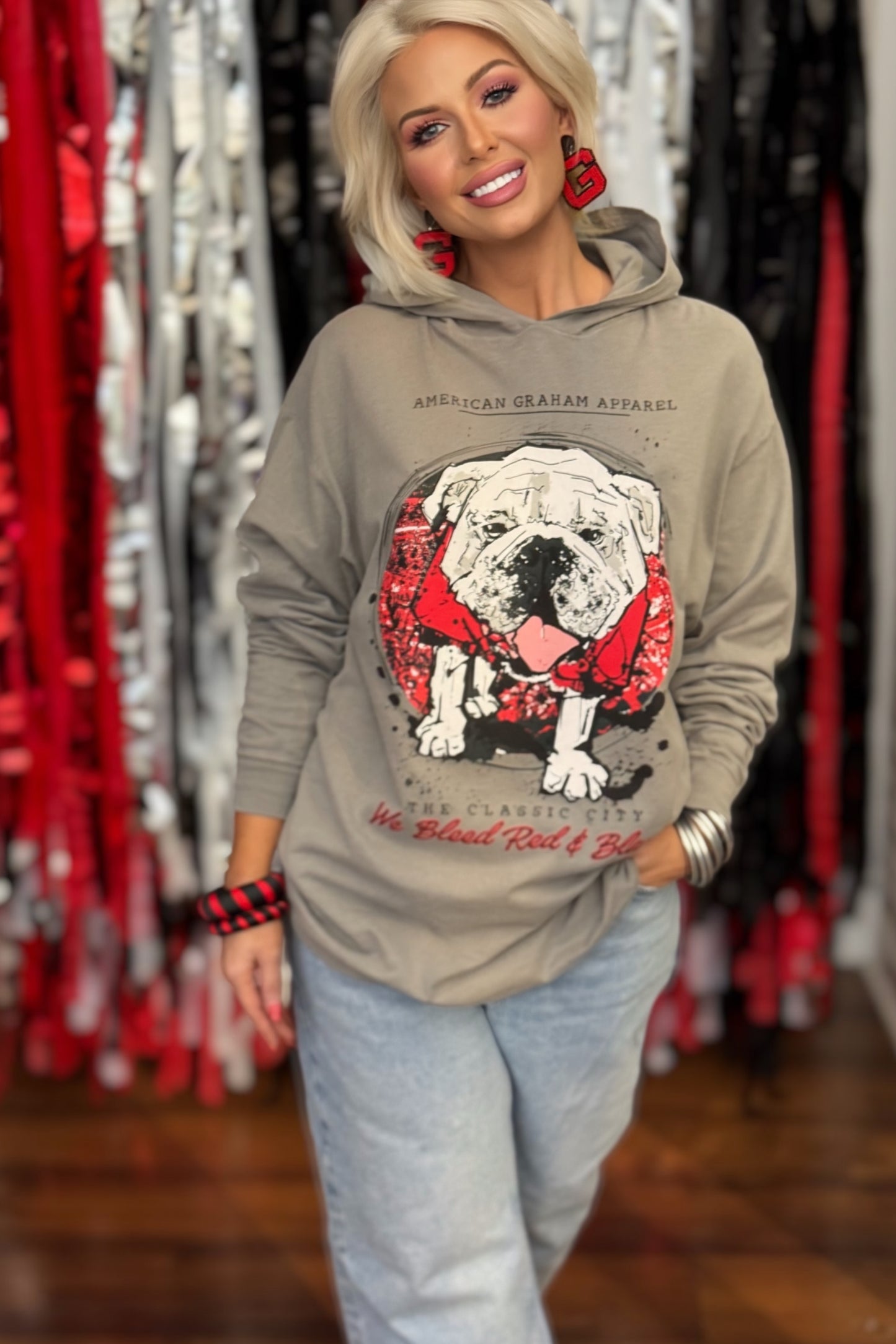 A person wearing a gray hooded long sleeve t-shirt with a large graphic print of a bulldog wearing a red bandana, with the text 'AMERICAN GRAHAM APPAREL' and 'The Classic City We Blood Red & Black' on the front.