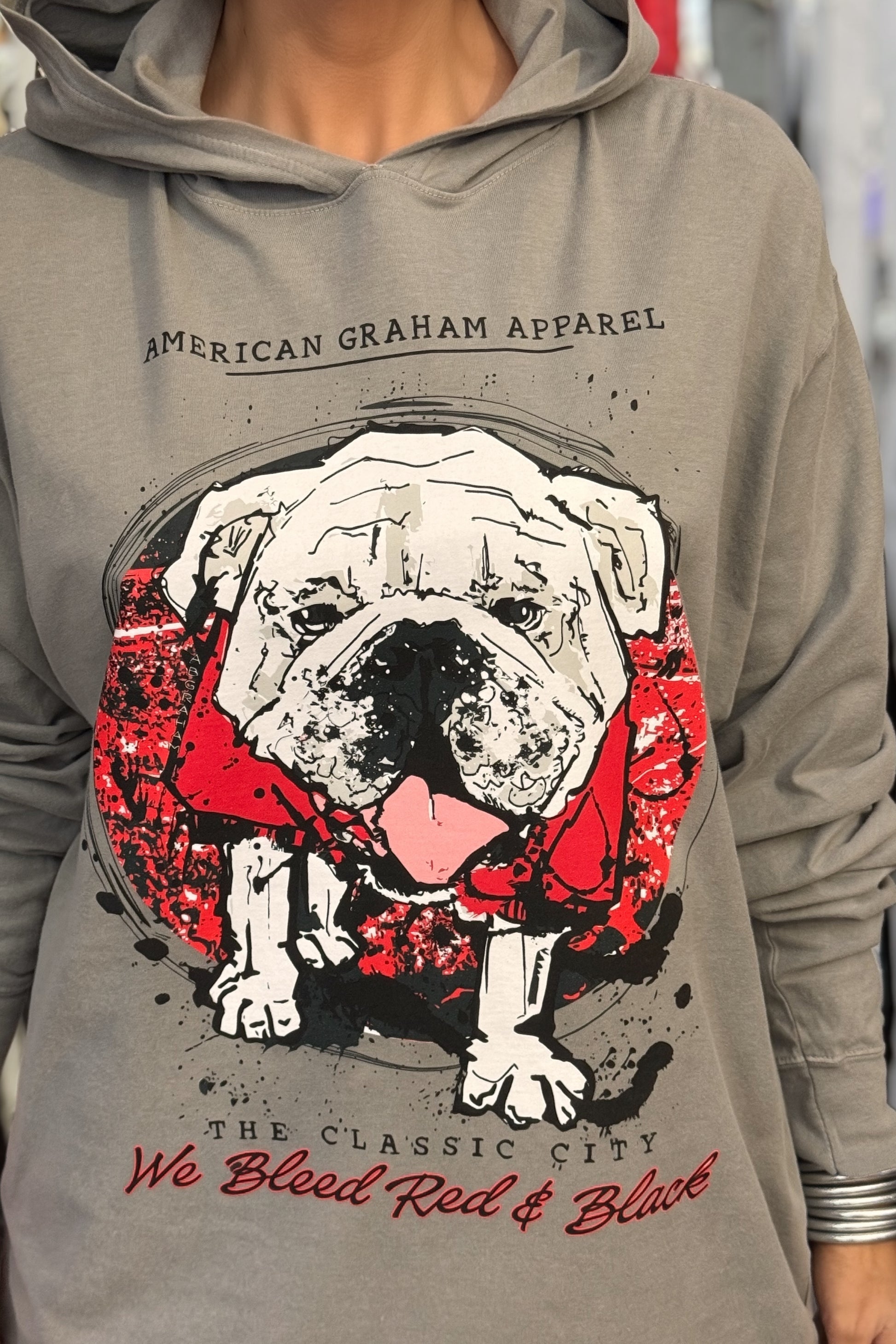 A person wearing a gray hooded long sleeve t-shirt with a large graphic print of a bulldog wearing a red bandana, with the text 'AMERICAN GRAHAM APPAREL' and 'The Classic City We Blood Red & Black' on the front.