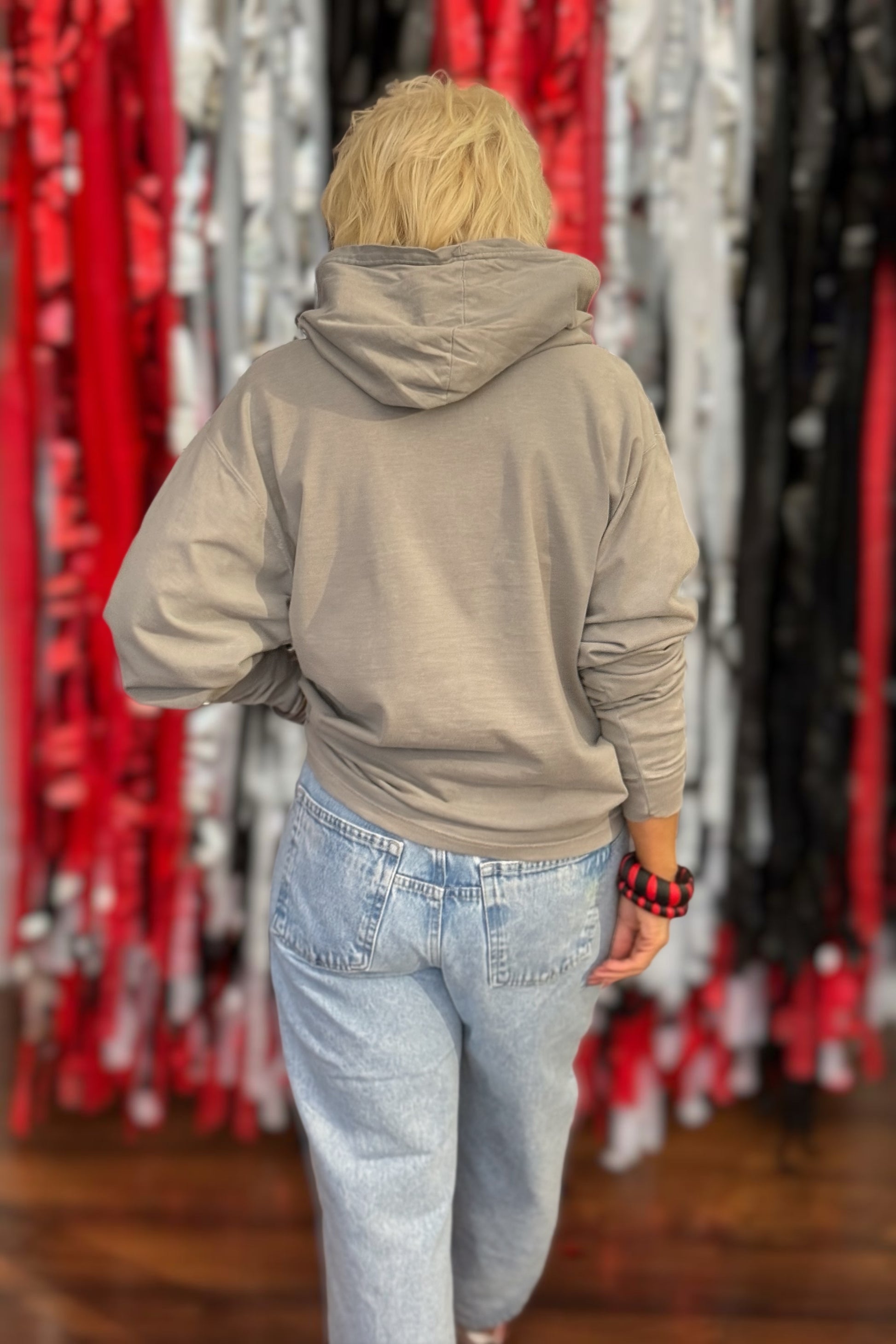 A person viewed from behind wearing a light taupe-beige pullover hoodie and light-wash blue jeans, standing in front of a display of colorful hanging tinsel garlands.