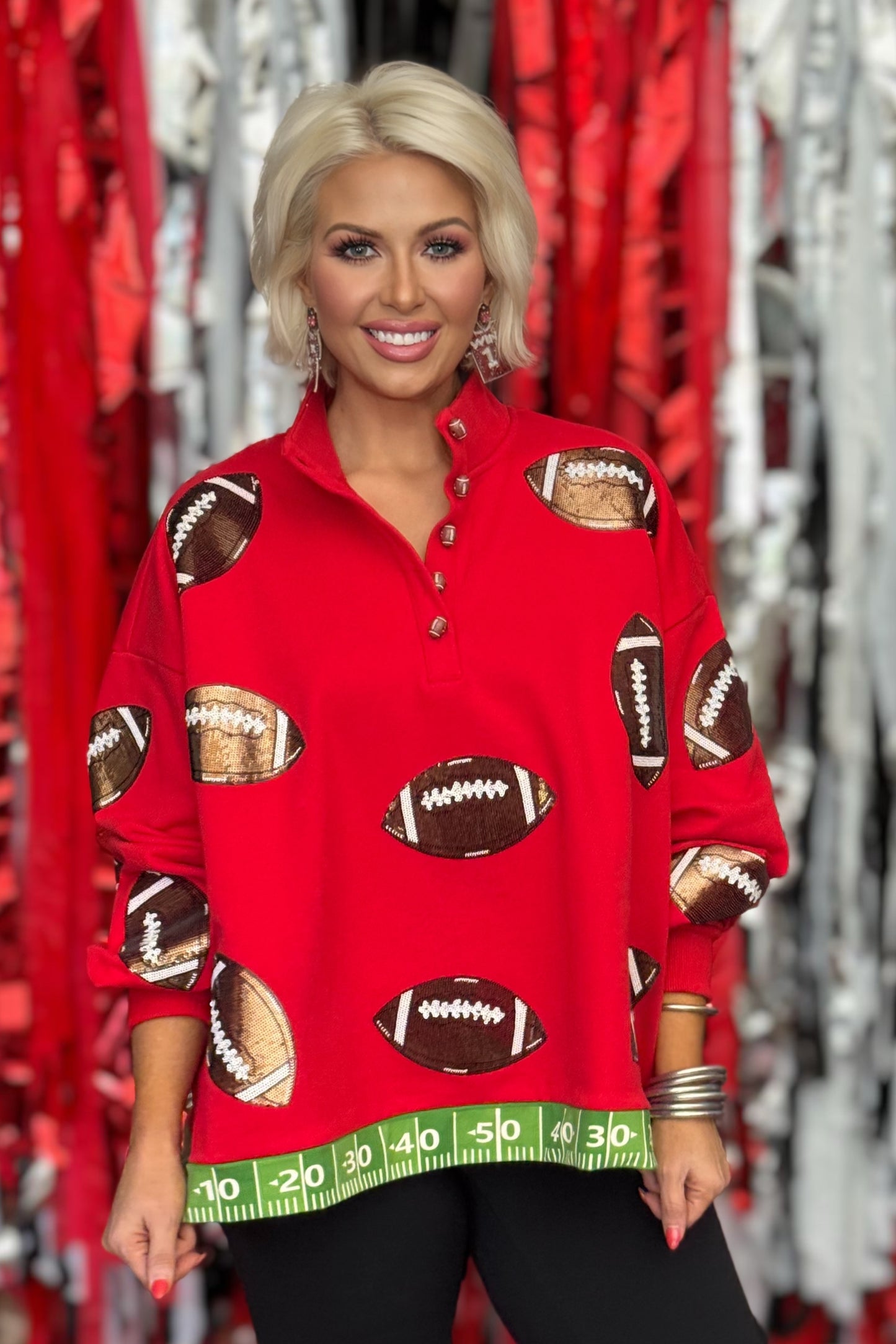 Woman wearing an oversized red game-day pullover with all-over brown and bronze sequin football appliqués and green football field hem trim.